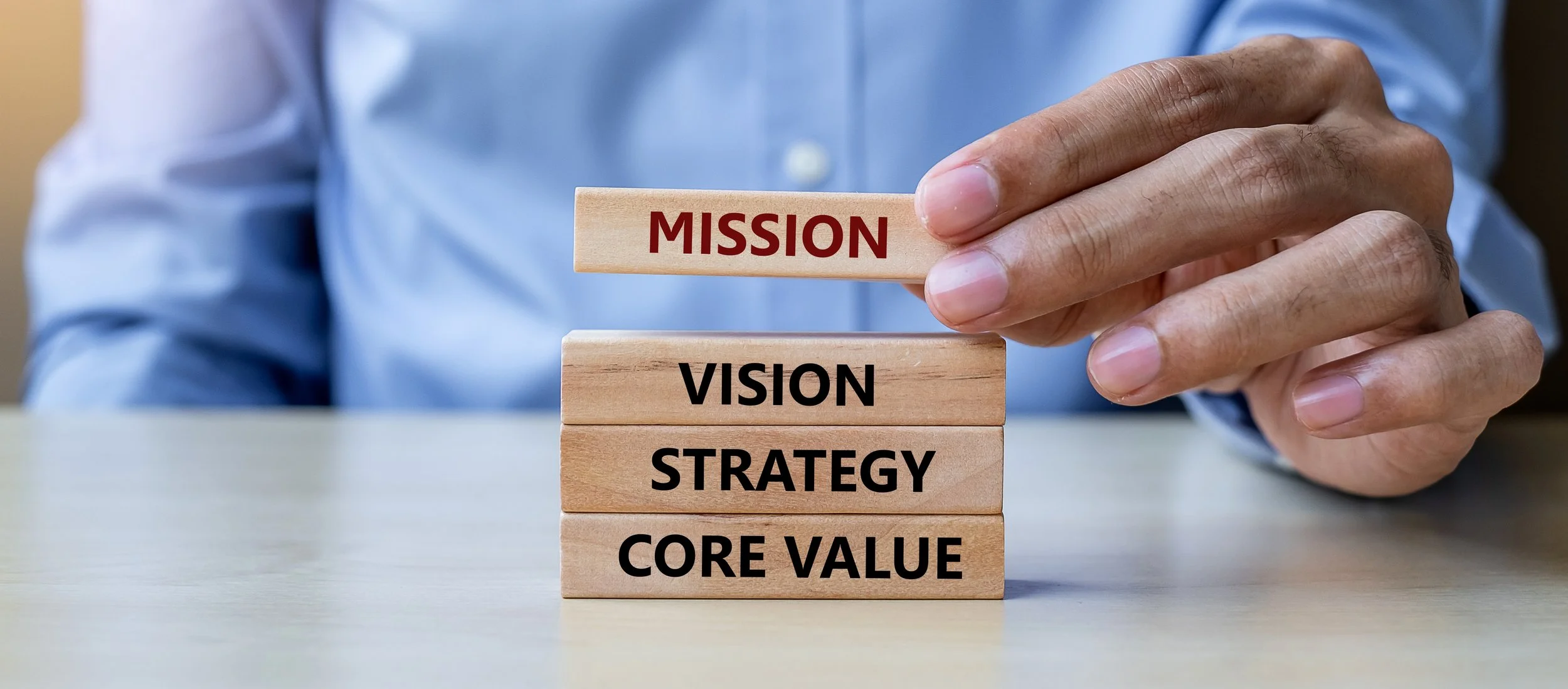 A person in a blue shirt places a wooden block labeled 'MISSION' on top of four stacked blocks labeled 'VISION,' 'STRATEGY,' and 'CORE VALUE.'