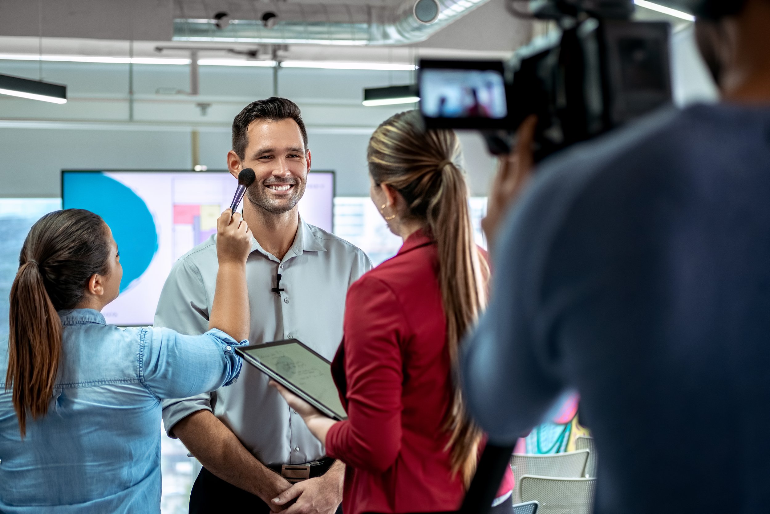 A man is being filmed as he is getting his makeup done by a woman holding a makeup brush, while other women are taking notes and filming him in a modern office setting.