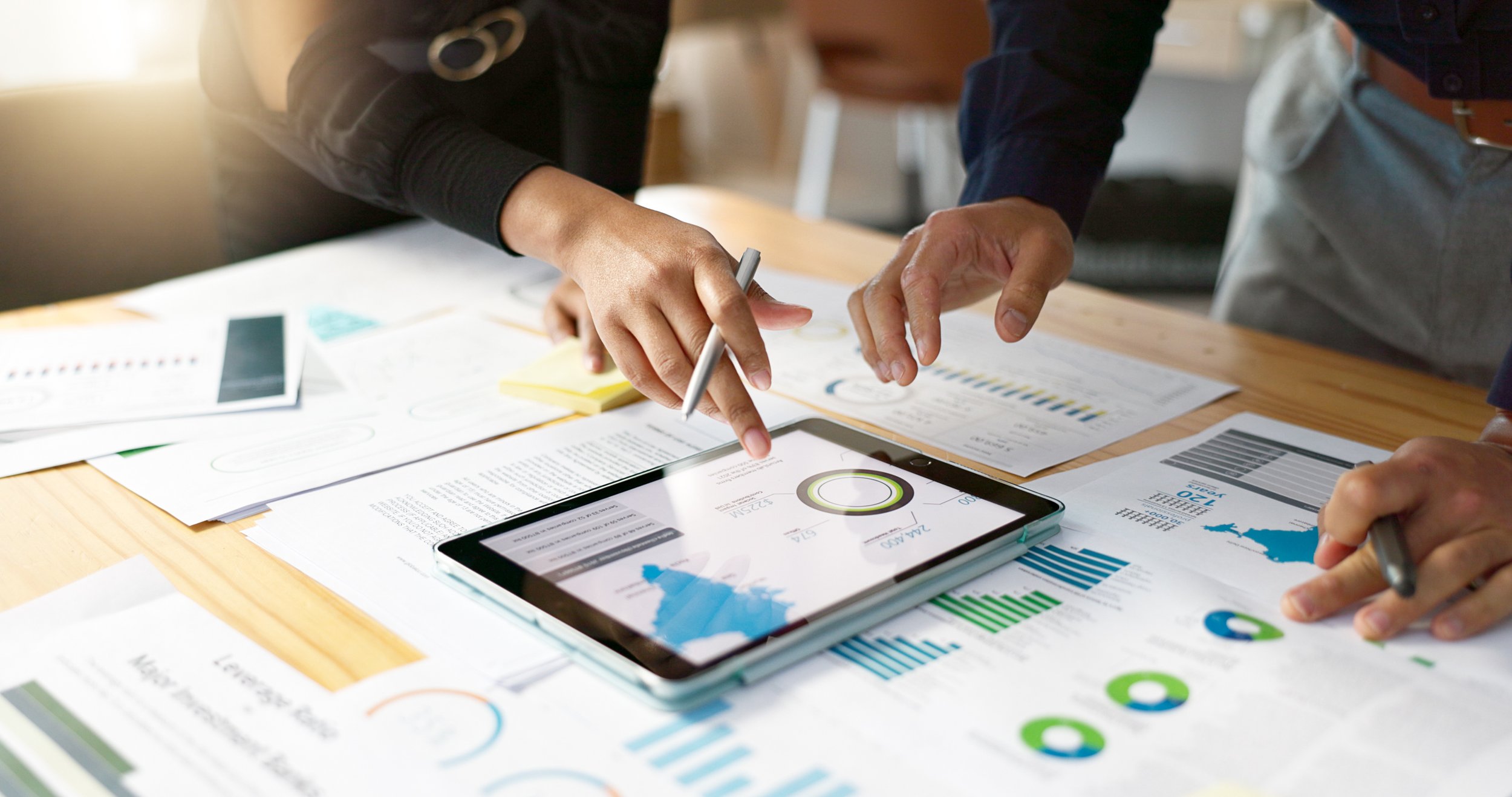 People reviewing documents and data charts on a tablet and paper spread on a wooden table.
