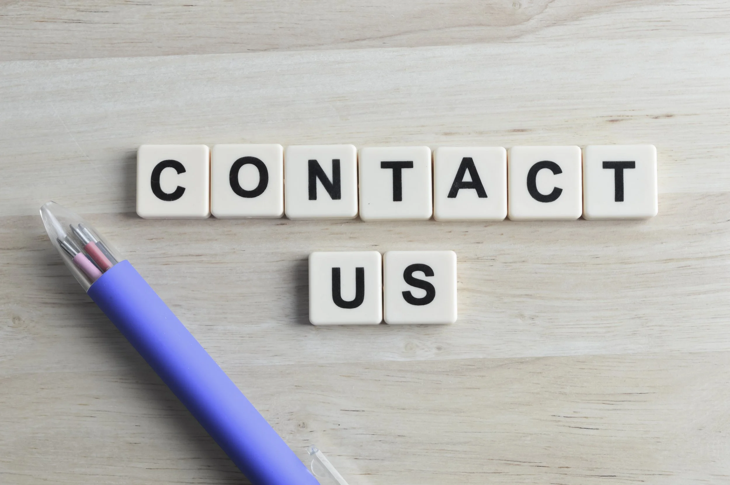 Scrabble tiles spell out 'CONTACT US' on a light-colored wooden surface, with a light purple pen with three small red, pink, and purple clips placed on the lower left side.