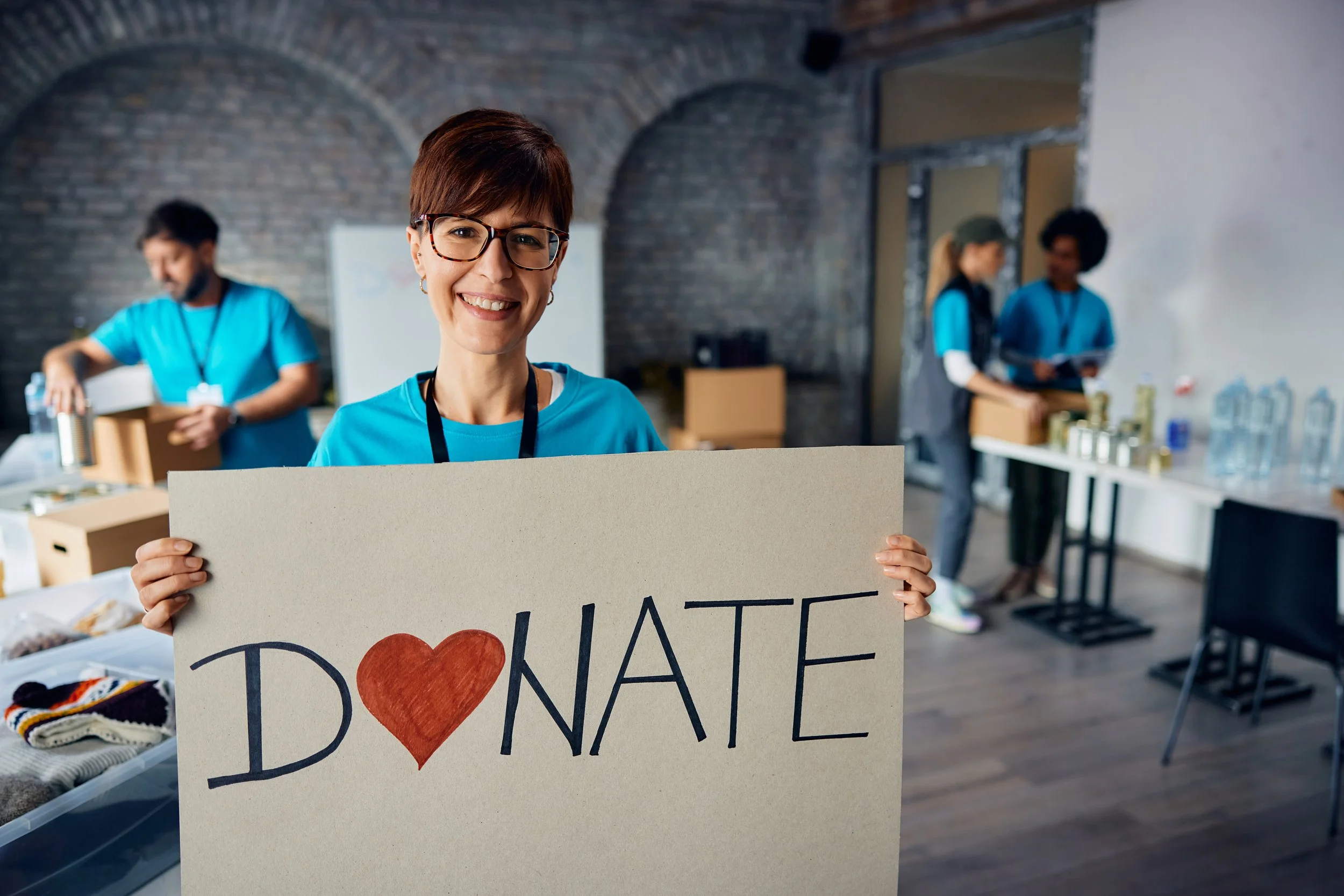 Woman smiling and holding a handmade sign that reads 'I ❤️ NATE' at a donation event, with other volunteers in the background organizing items.