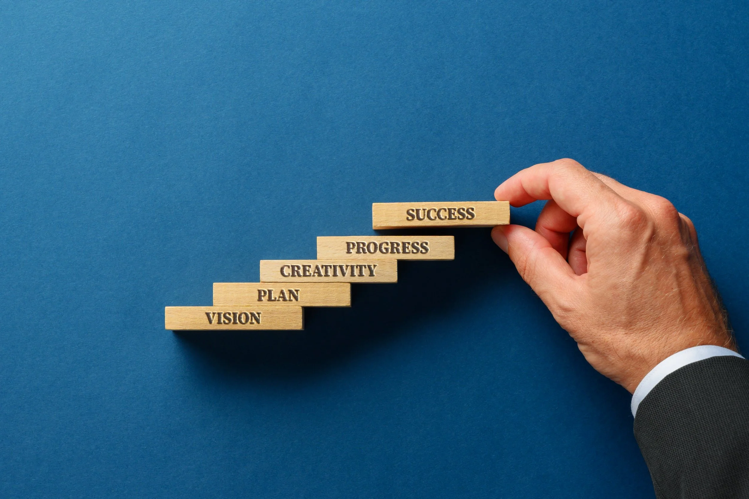 Hand placing a wooden block labeled 'SUCCESS' on a staircase made of other wooden blocks labeled 'VISION', 'PLAN', 'CREATIVITY', 'PROGRESS', which are arranged upward on a blue background