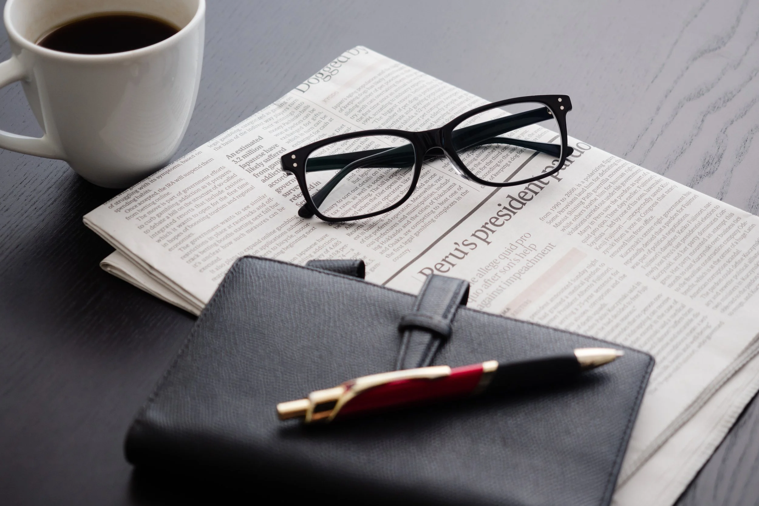 A cup of black coffee, black eyeglasses, a newspaper, a closed black notebook, and a pen on a dark wooden surface.
