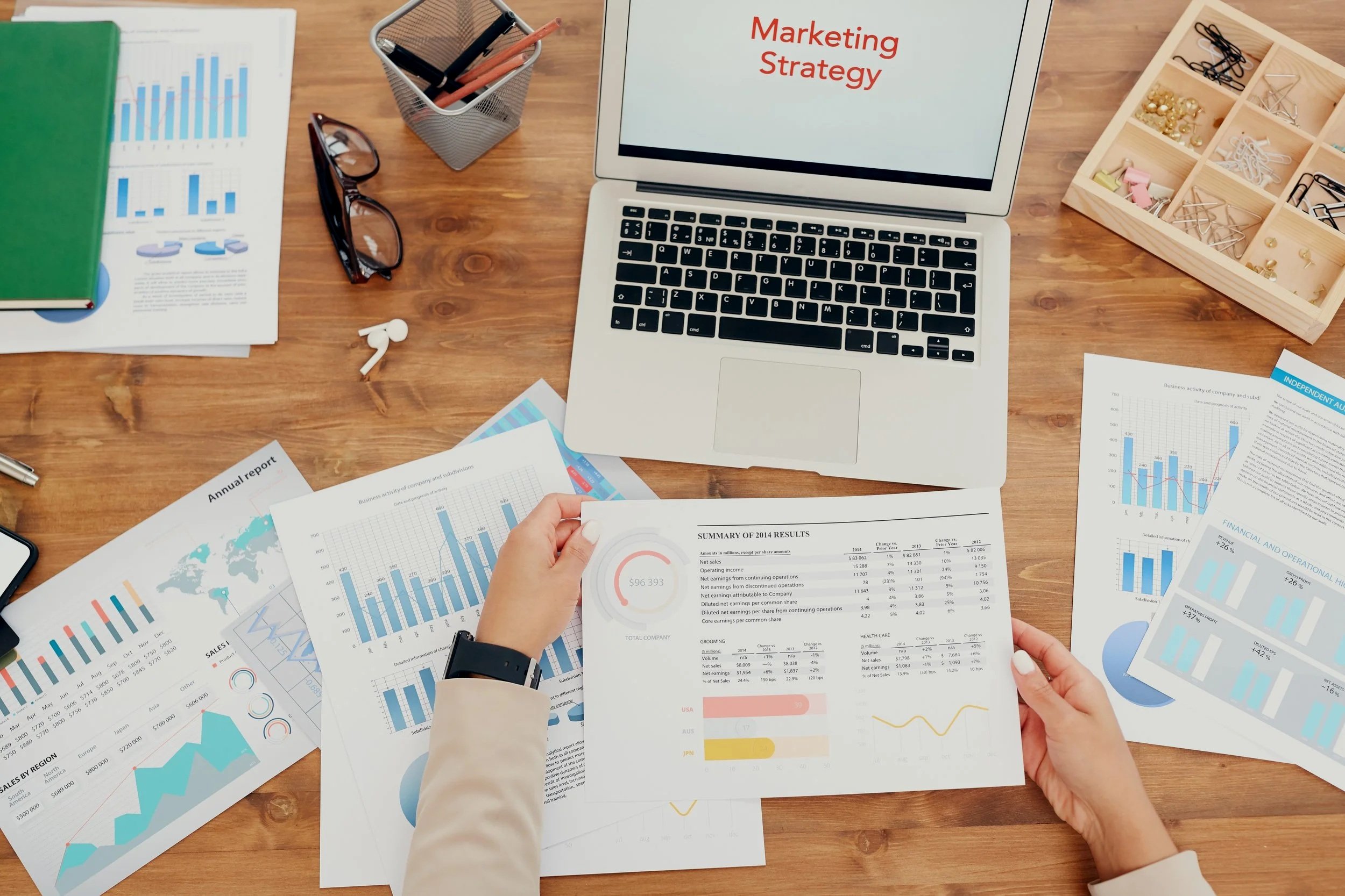 Work desk with financial reports, graphs, and charts, a laptop displaying 'Marketing Strategy,' eyeglasses, a notebook, and office supplies, indicating a business or marketing planning session.