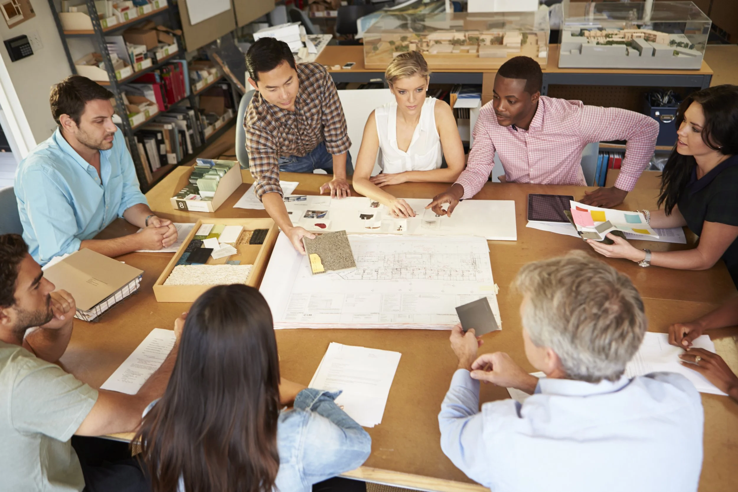 Group of diverse professionals gathered around a table during a meeting, discussing architectural plans and samples in an office setting.