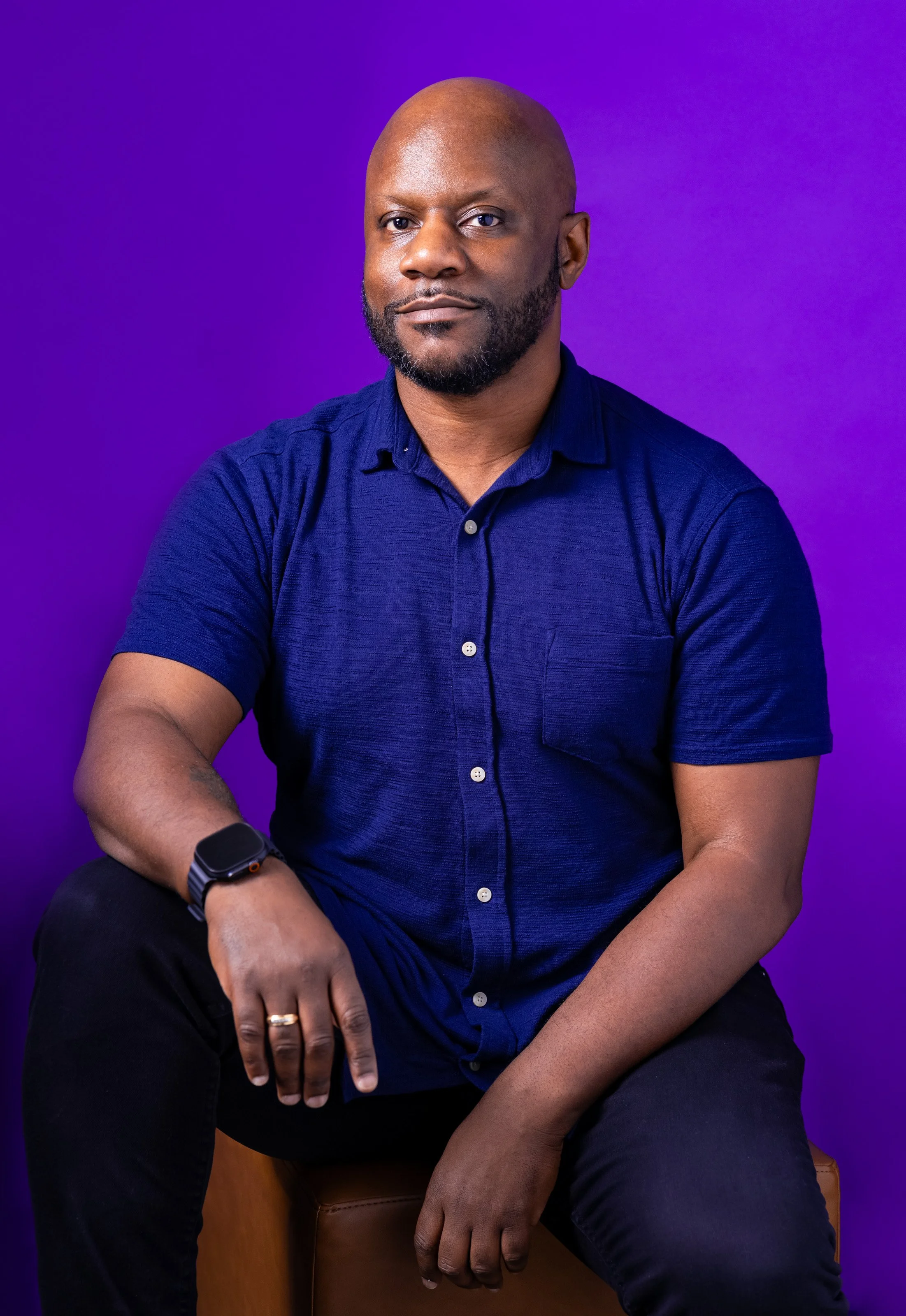 A man with a beard and shaved head wearing a navy blue shirt seated on a brown leather stool against a purple backdrop.