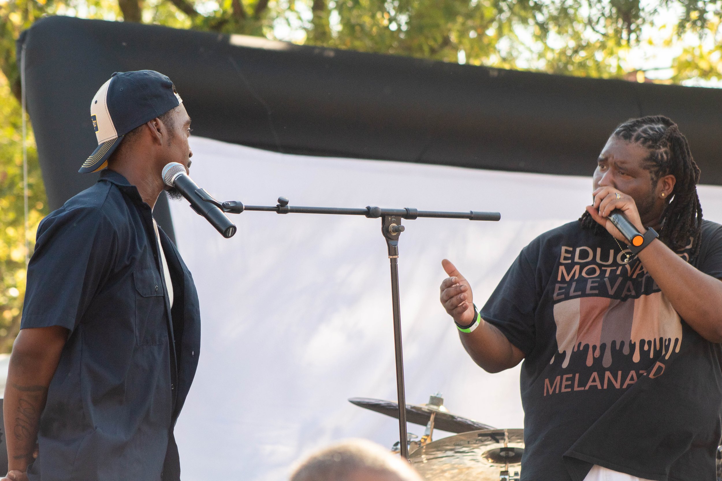 Two men are having a conversation outdoors at a music or speech event, with one holding a microphone and the other standing near a music stand, wearing a hat and casual clothing.