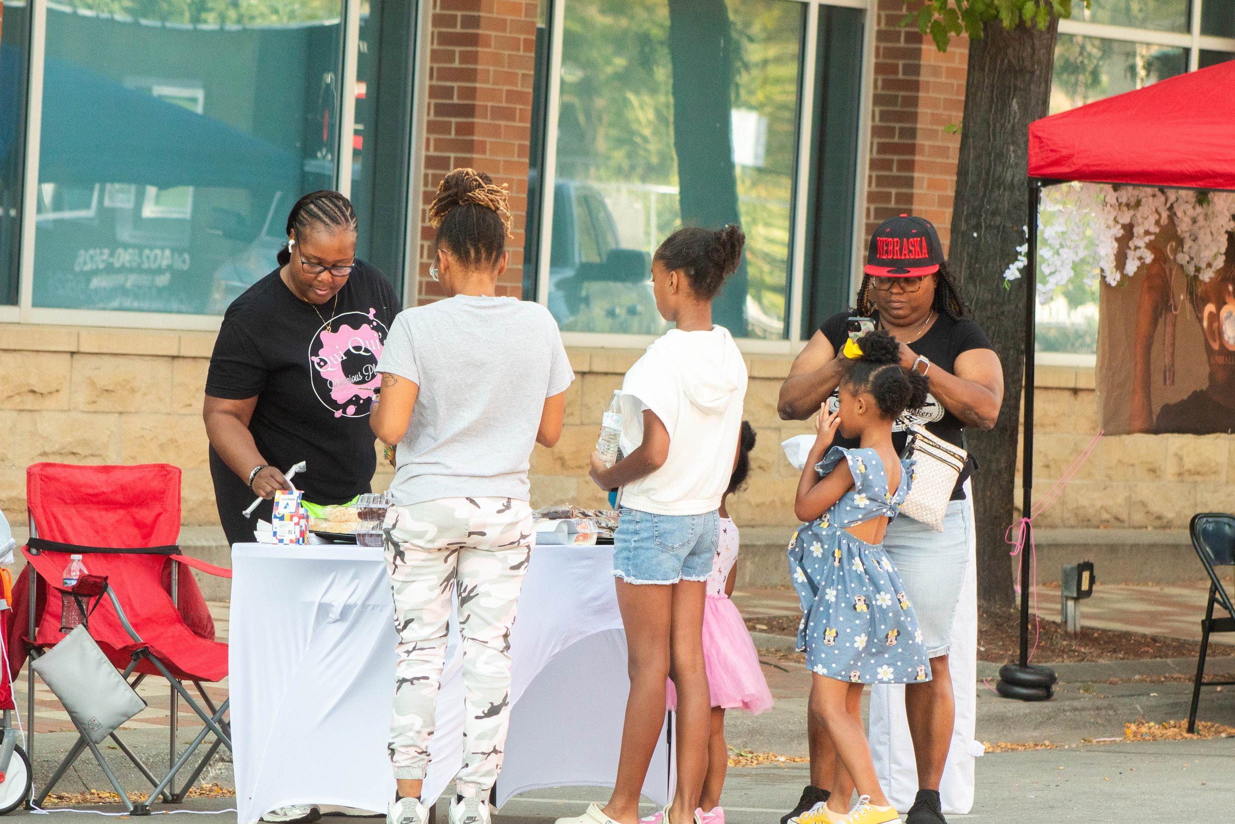A group of people, mostly children and women, gathered around a table at an outdoor event. The women and children are engaging with items on the table. One woman is looking at her phone, while others are interacting or waiting in line. There is a red