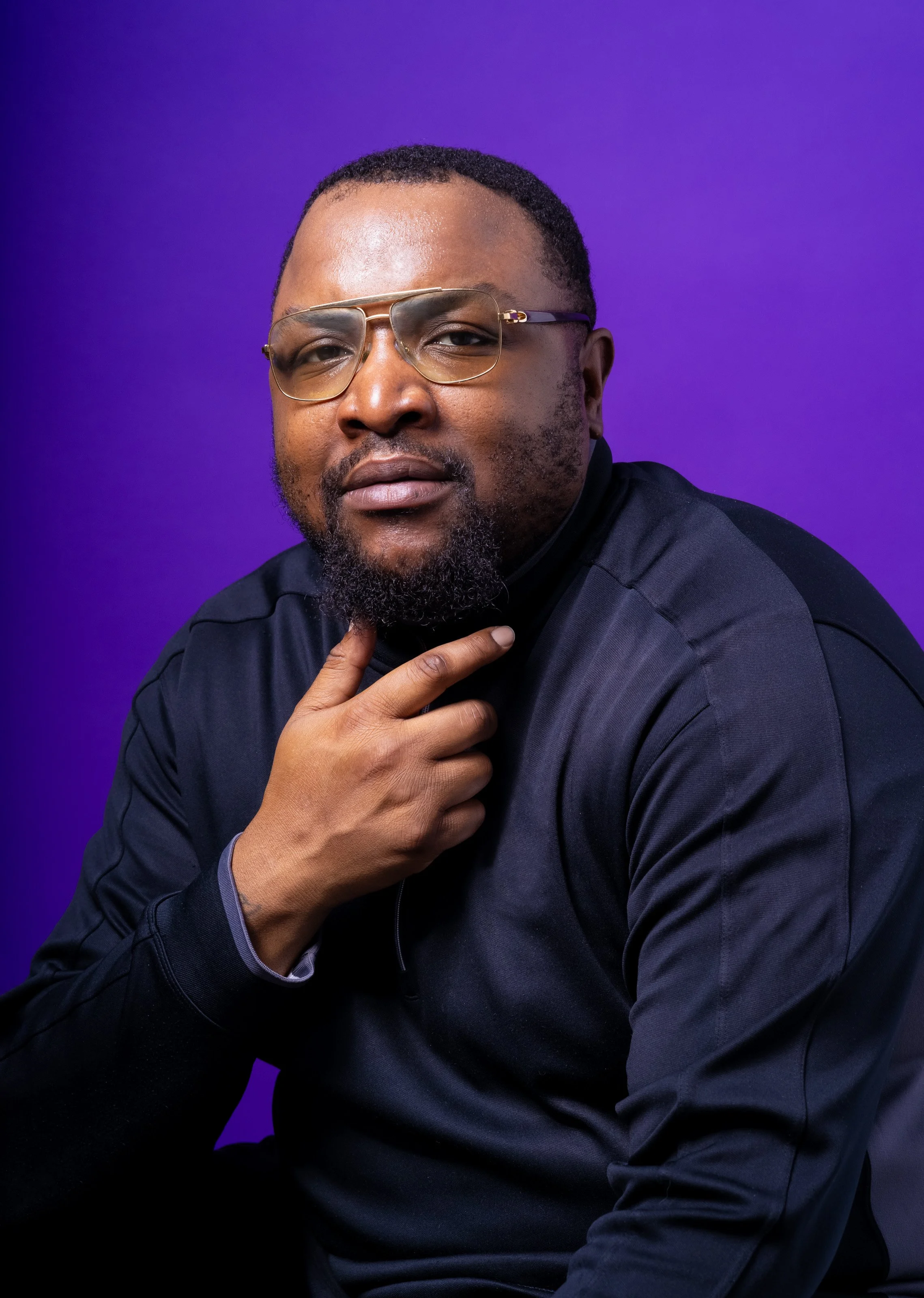 Portrait of a man with short hair, beard, and glasses, wearing a black jacket, against a purple background.