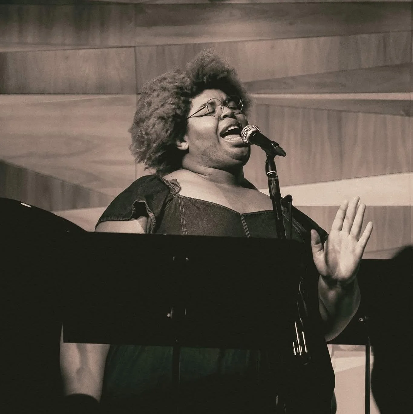 A woman with curly hair and glasses singing into a microphone, with her hand raised in a gesture of expression, in a setting with wooden background panels.