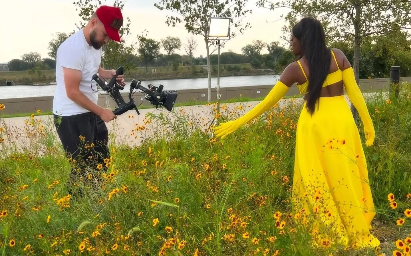 A woman in a bright yellow dress and yellow gloves is standing in a field of yellow and orange flowers near a river. A man with a camera stabilizer is filming her.