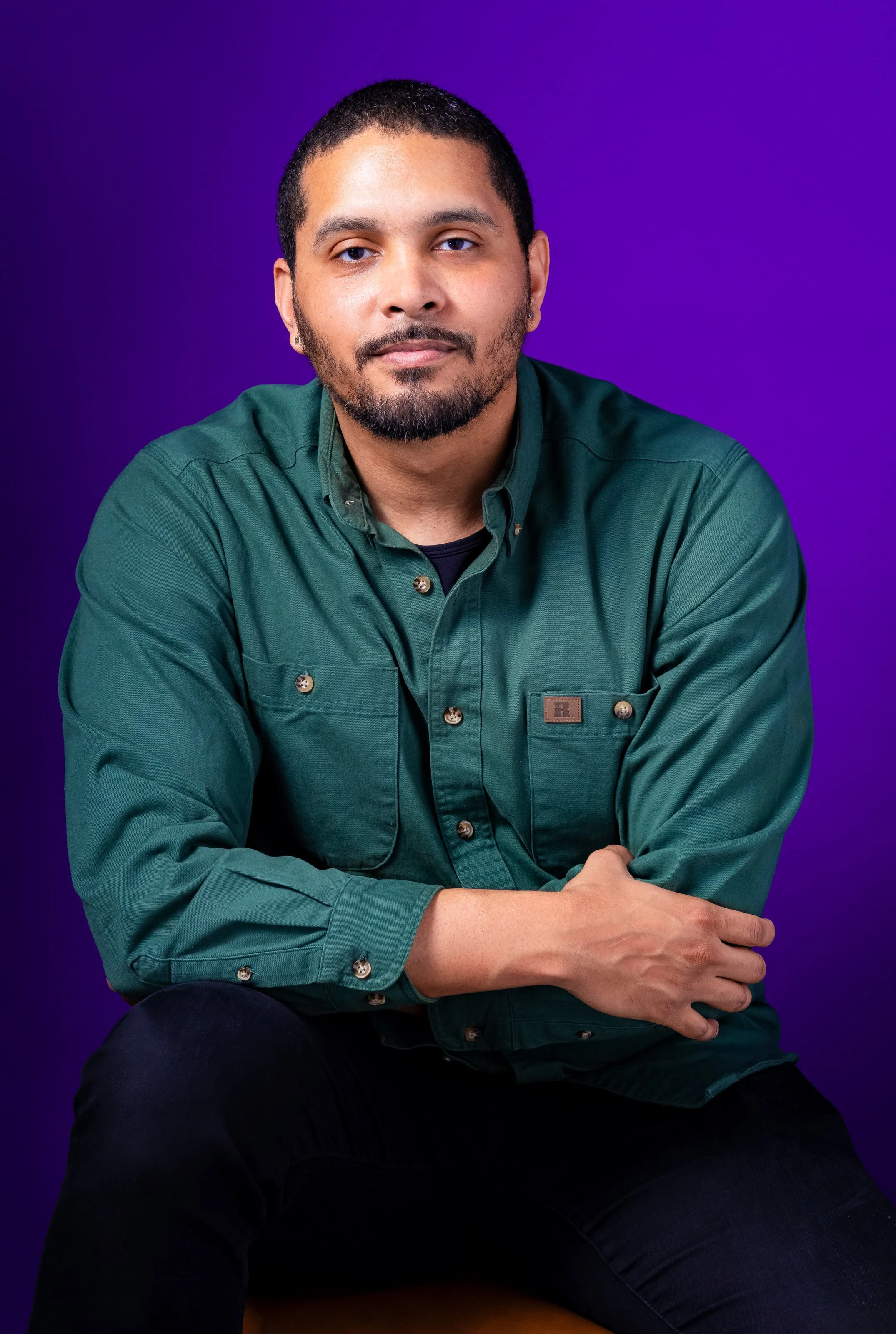 Portrait of a man with short dark hair, a beard, and wearing a green button-up shirt, sitting against a purple background.