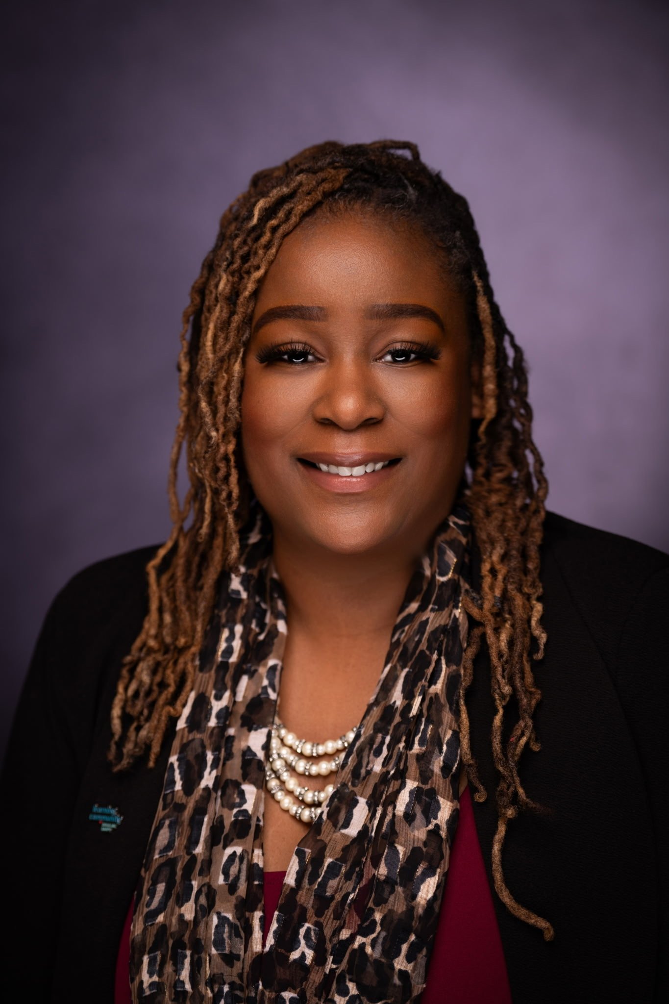 A woman with long, styled dreadlocks, smiling, wearing a black blazer, leopard print scarf, a pearl necklace, and a red top, against a purple background.