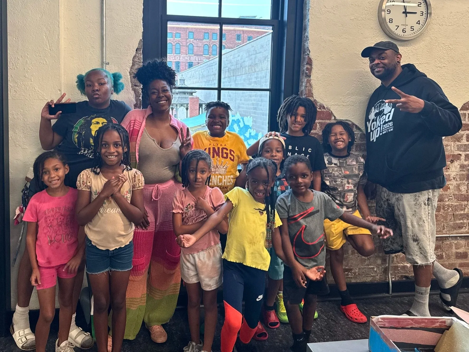 Group of children and two adults posing for a photo indoors near a large window. The children are smiling and making peace signs, and the adults are standing behind them, smiling. The background shows an urban building through the window, and there is a clock on the wall showing 12:05.