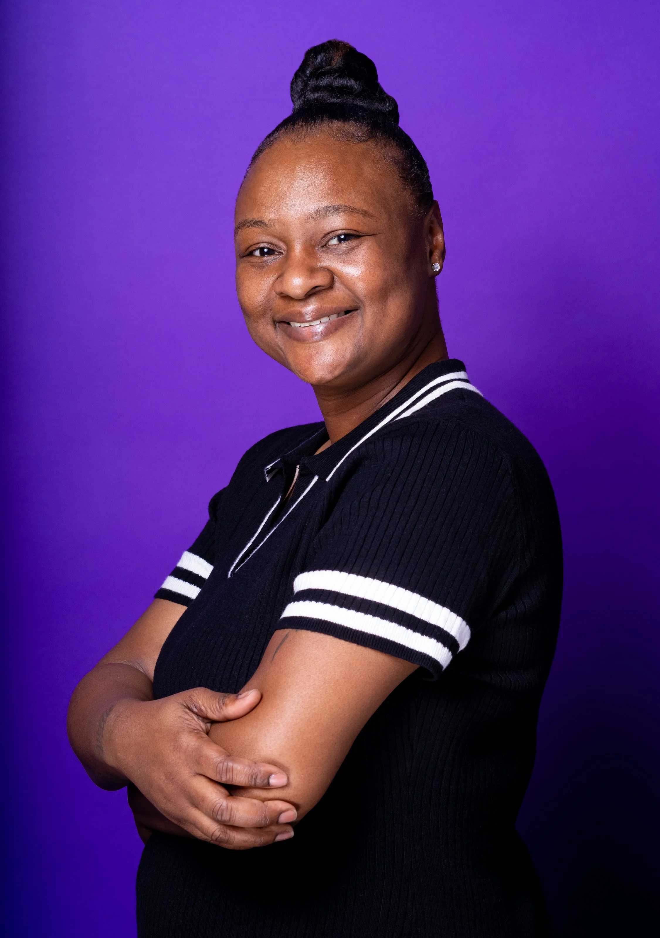 A smiling woman with a top knot hairstyle, wearing a black shirt with white stripes on the sleeves, standing against a purple gradient background.