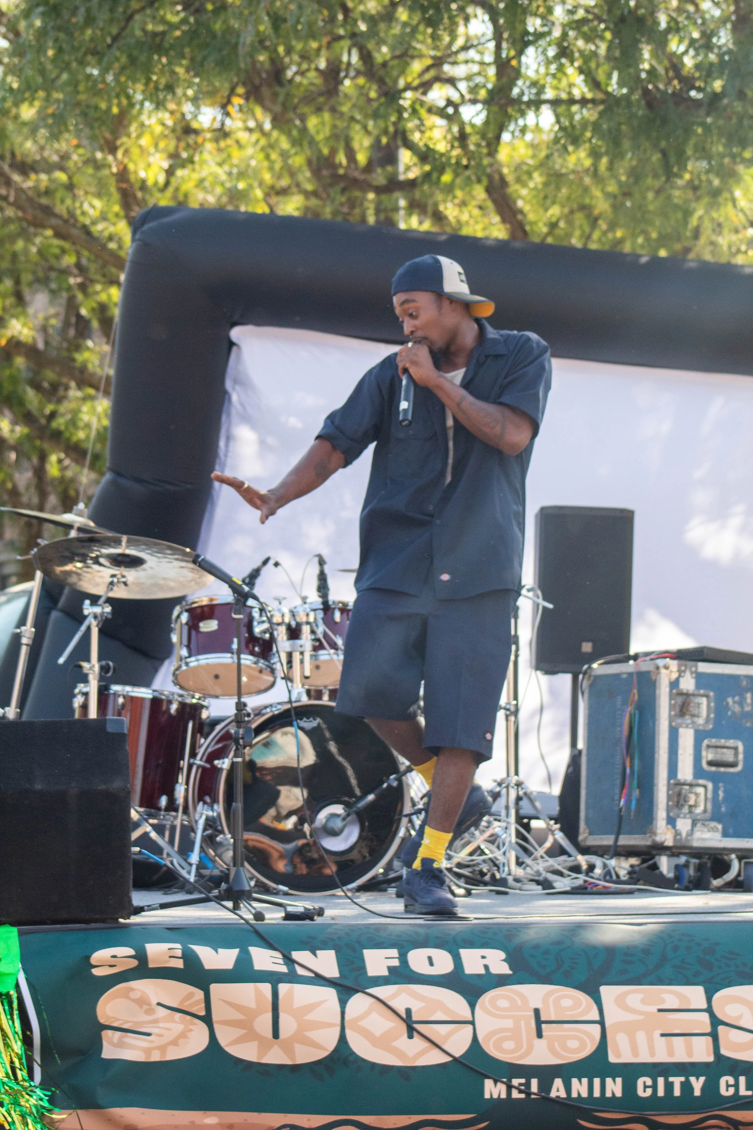 A man wearing a backwards cap, a dark shirt, and shorts performing with a microphone on an outdoor stage with musical equipment, surrounded by trees and a banner that reads "SEVEN FOR MELANIN CITY CLASSIC."