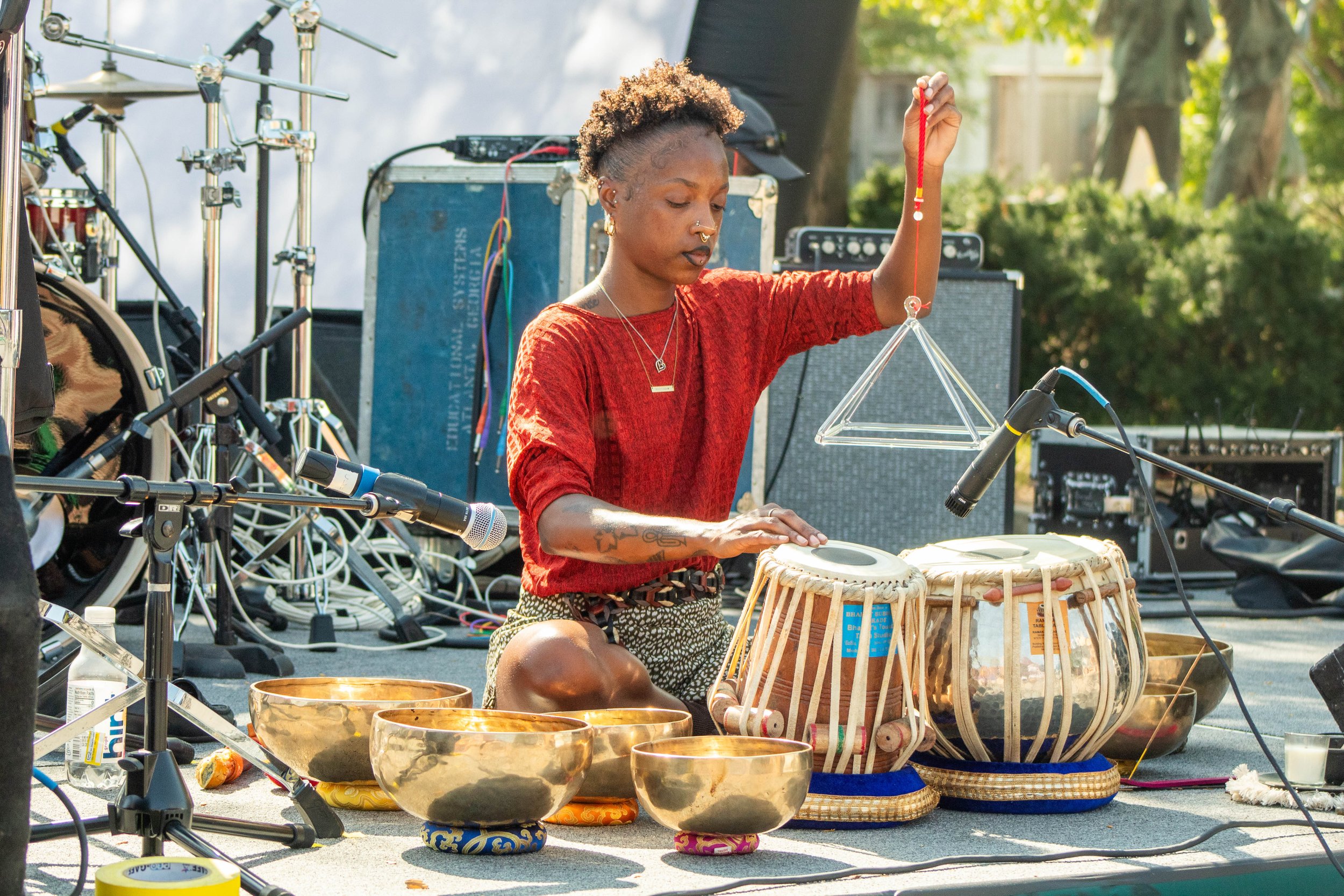 A woman with short curly hair, wearing a red top, playing a gong and a drum set outdoors during the daytime. She is surrounded by musical equipment and percussion instruments, with sunlight filtering through green trees in the background.