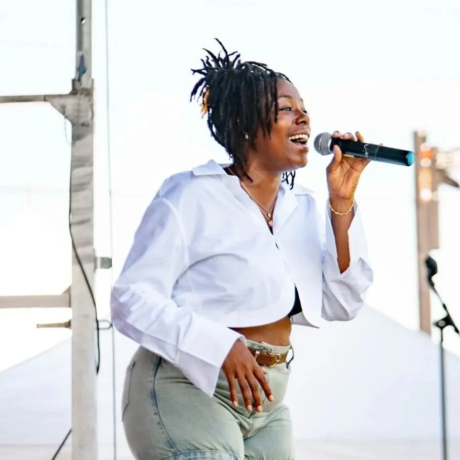 A woman with dreadlocks singing into a microphone on stage, wearing a white cropped shirt and light-colored pants.