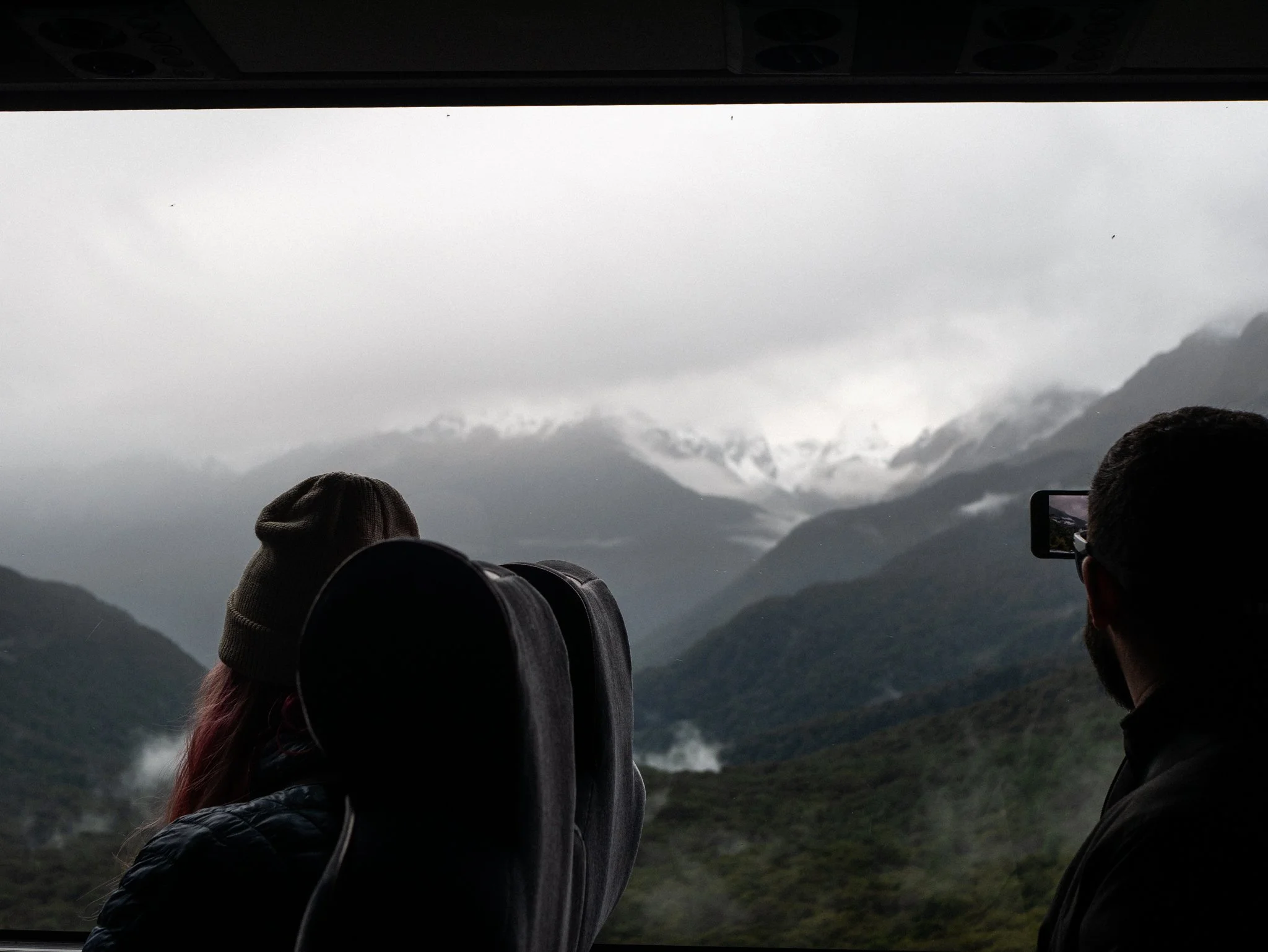 Two people looking out a bus window at a cloudy, mountainous landscape with mist.