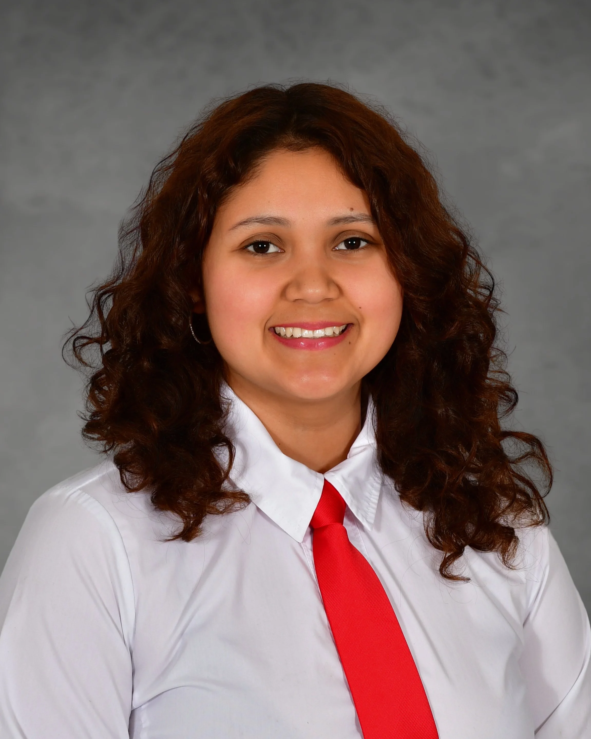 Portrait of a smiling woman with curly brown hair wearing a white shirt, standing in front of a brick wall.