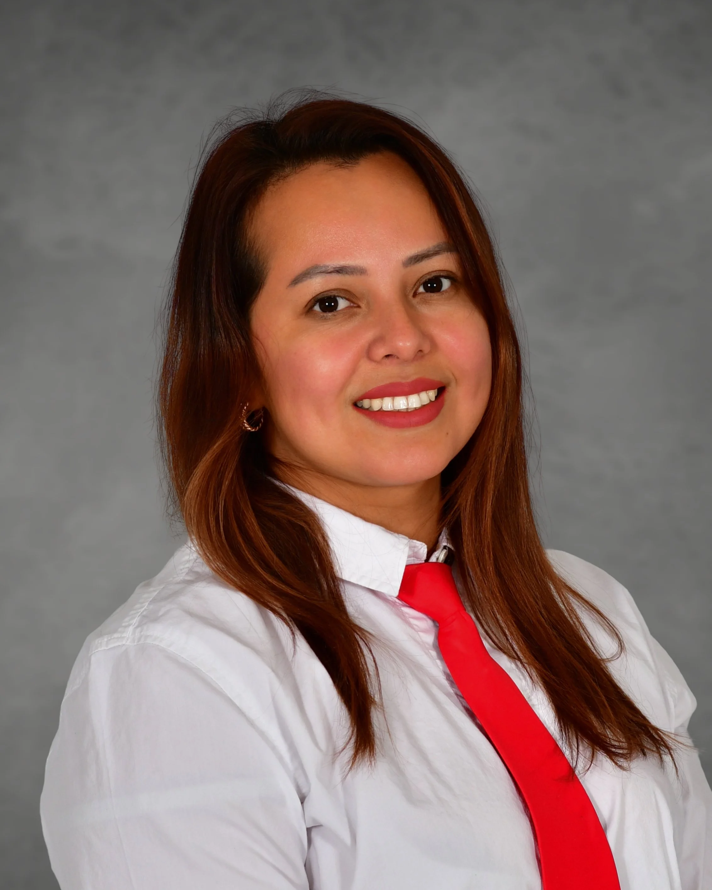 A woman with long, reddish-brown hair, wearing a white button-up shirt, standing against a red brick wall.