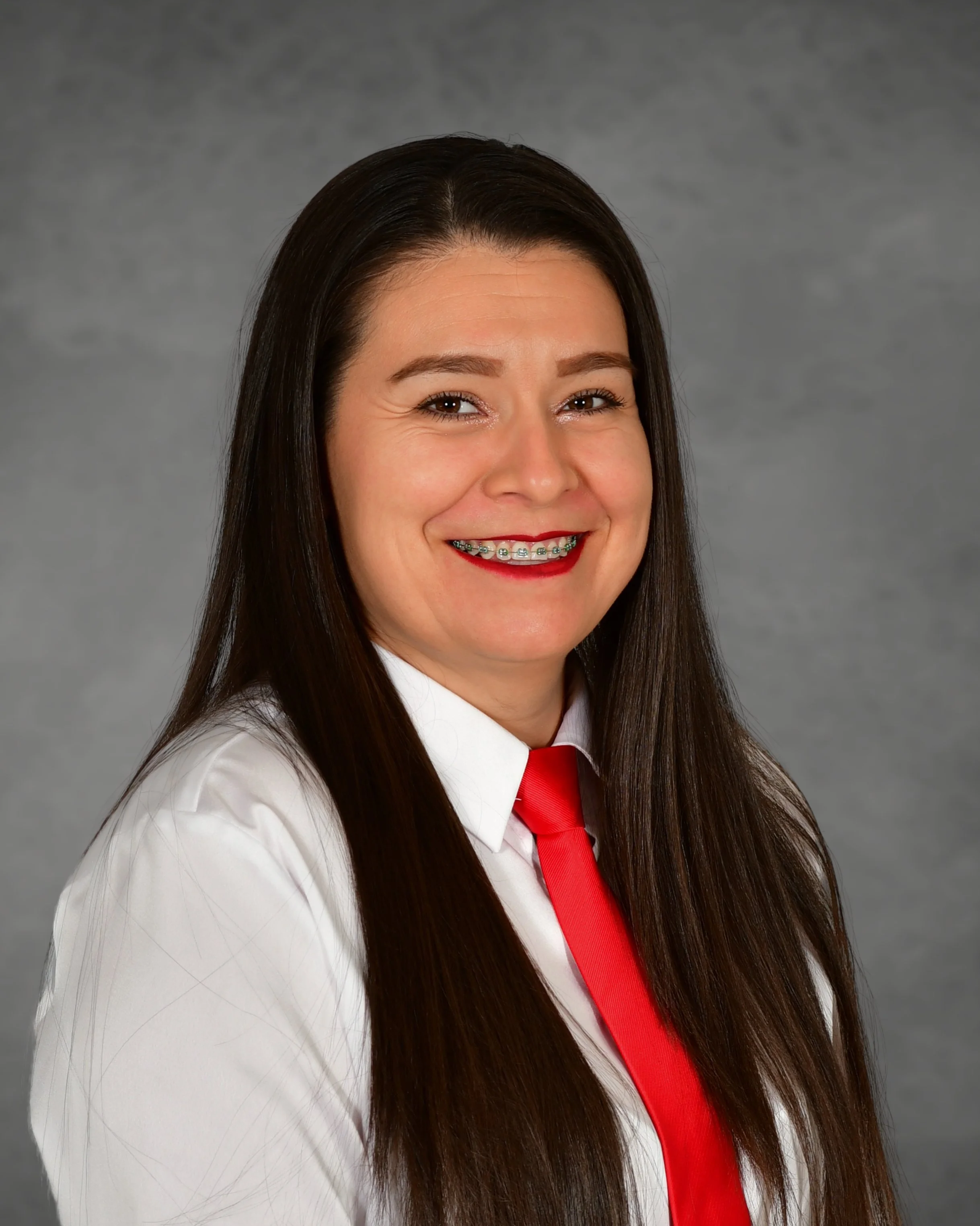 A woman with long dark hair wearing braces and a white shirt stands against a red brick wall, smiling.