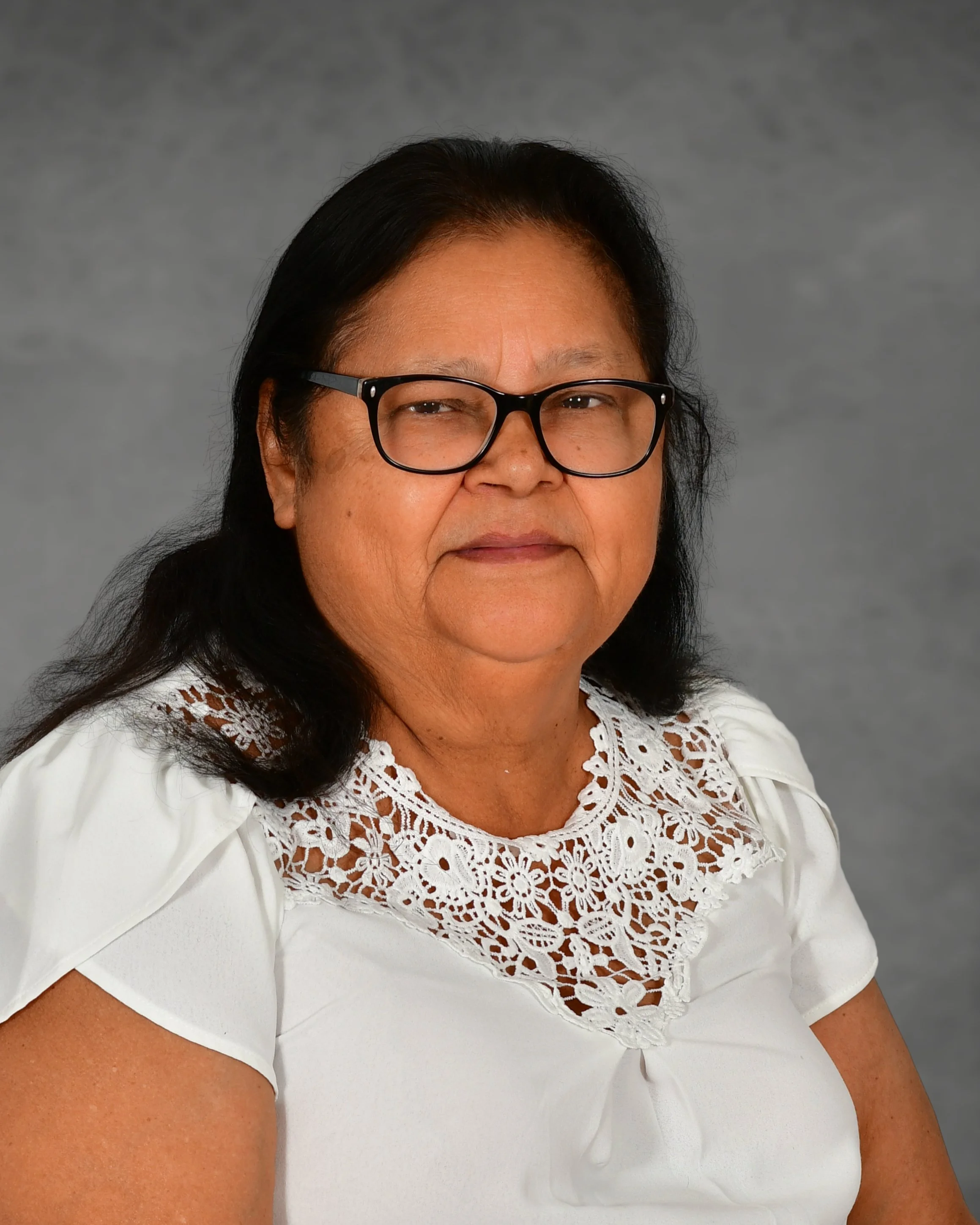 A woman with glasses and dark hair stands against a brick wall, smiling. She is wearing a white blouse with lace details and a gold necklace.