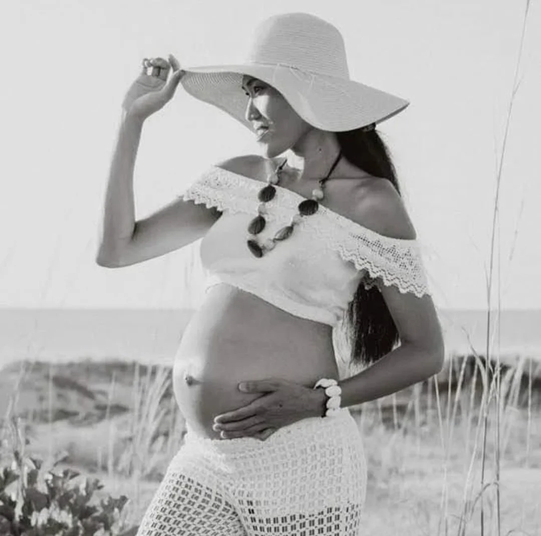 A pregnant woman in a crochet top, wearing a large sun hat, necklace, and bracelet, standing on a beach with grass in the background.