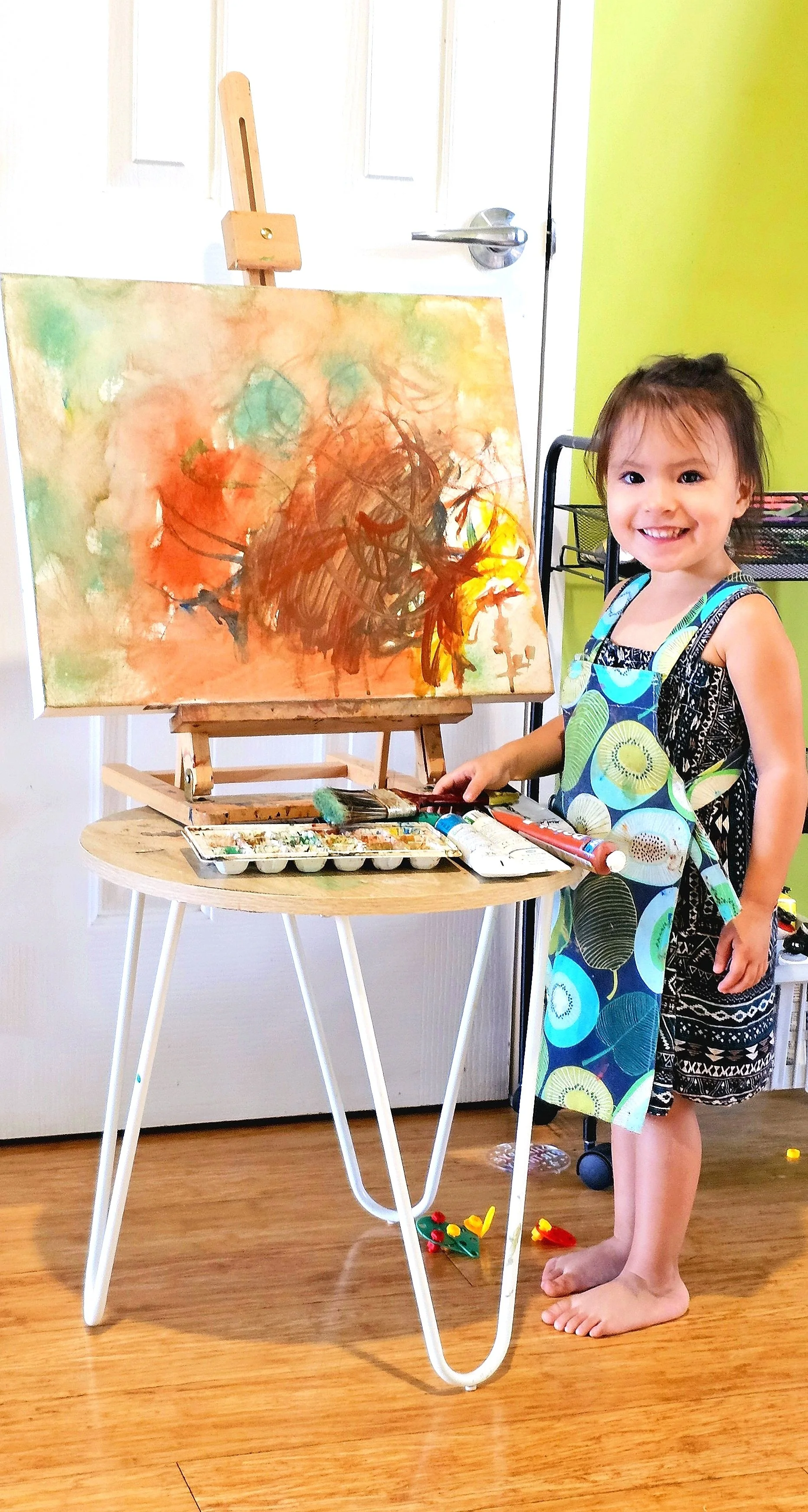 Child painting on an easel with an abstract artwork, wearing a colorful apron, standing on wooden floor.