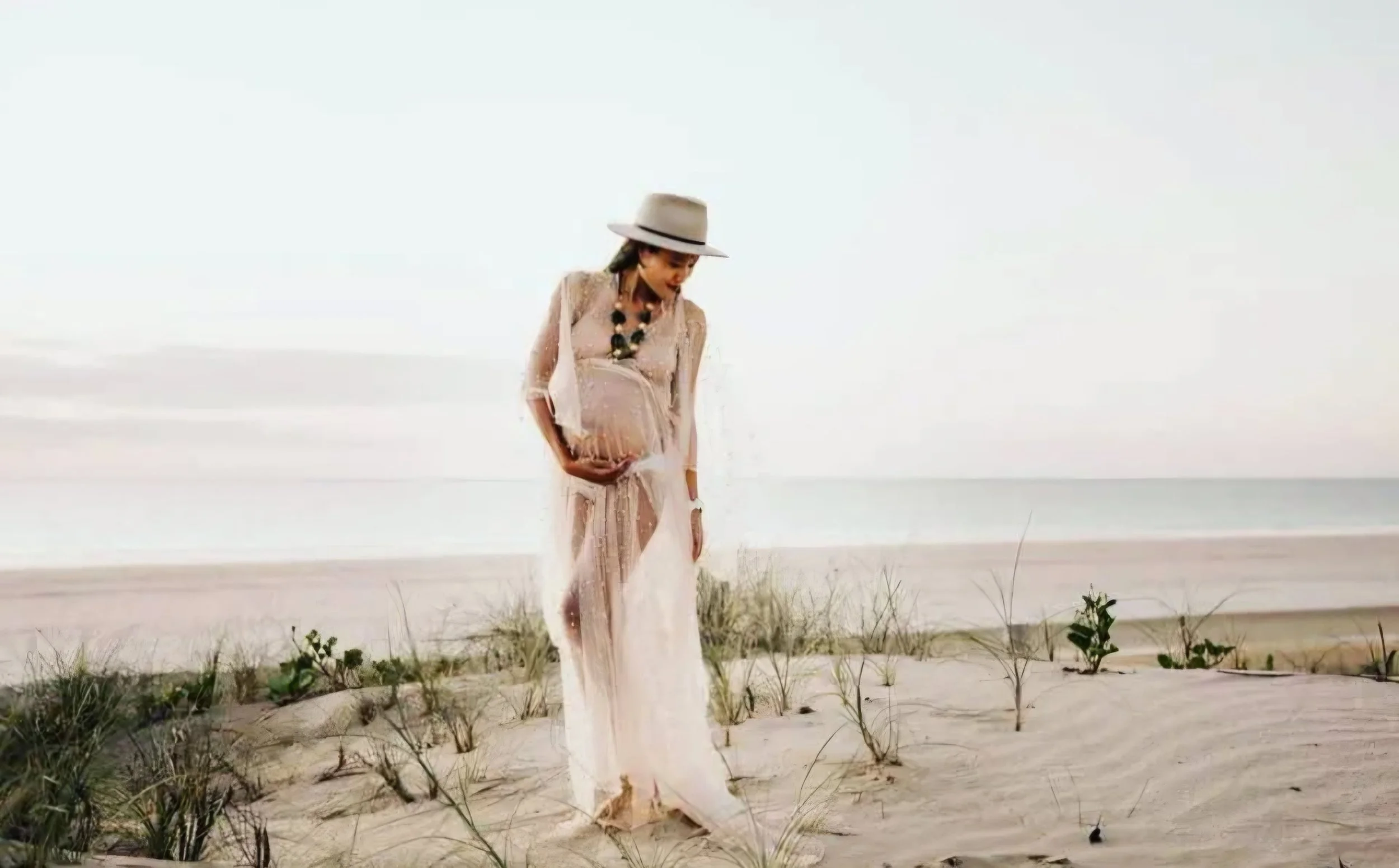A pregnant woman in a flowing dress and hat stands on the beach, gently holding her belly, with grass and ocean in the background.