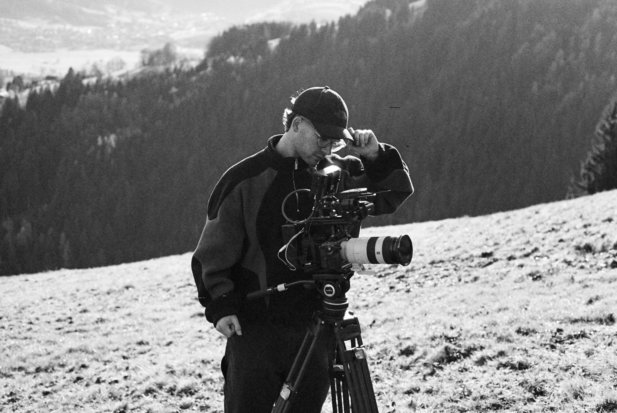Ein junger Mann steht mit einer Videokamera auf einem Stativ in einer bergigen, ländlichen Landschaft, schaut nach unten und trägt eine Brille, eine Mütze und eine Jacke, im Hintergrund sind Bäume und Hügel sichtbar.