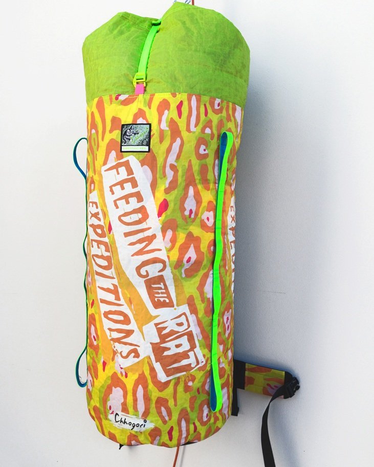 Colorful hiking or climbing gear bag with a leopard-print design, featuring text that reads "FEEDING THE REVOLUTION" and a small label with the brand name "Chogori." The bag has green and blue straps, and a black strap at the bottom.