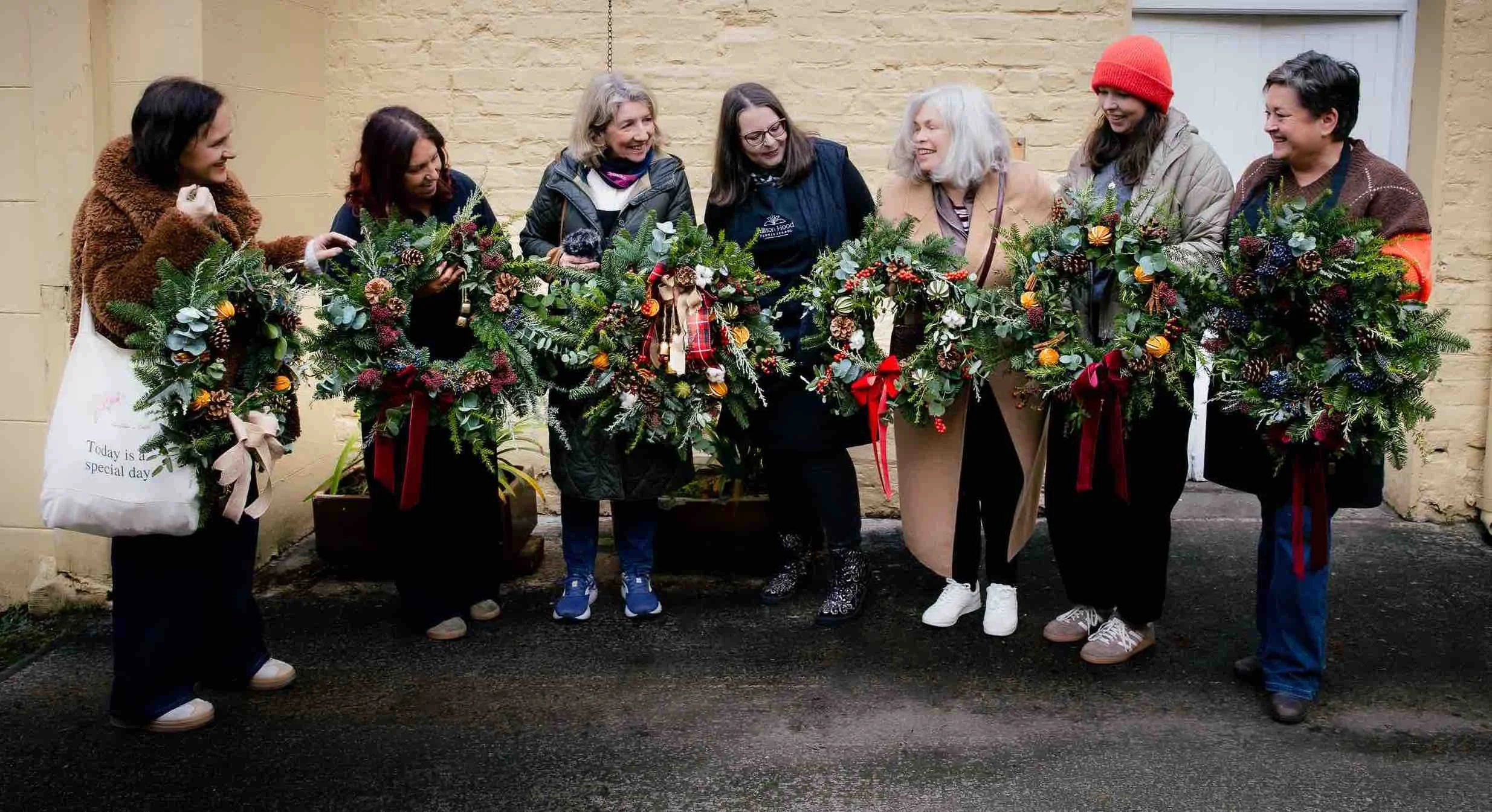 Group of seven women standing outdoors in front of a yellow brick wall, holding festive Christmas wreaths decorated with pine, berries, and ribbons, smiling and engaging with each other.