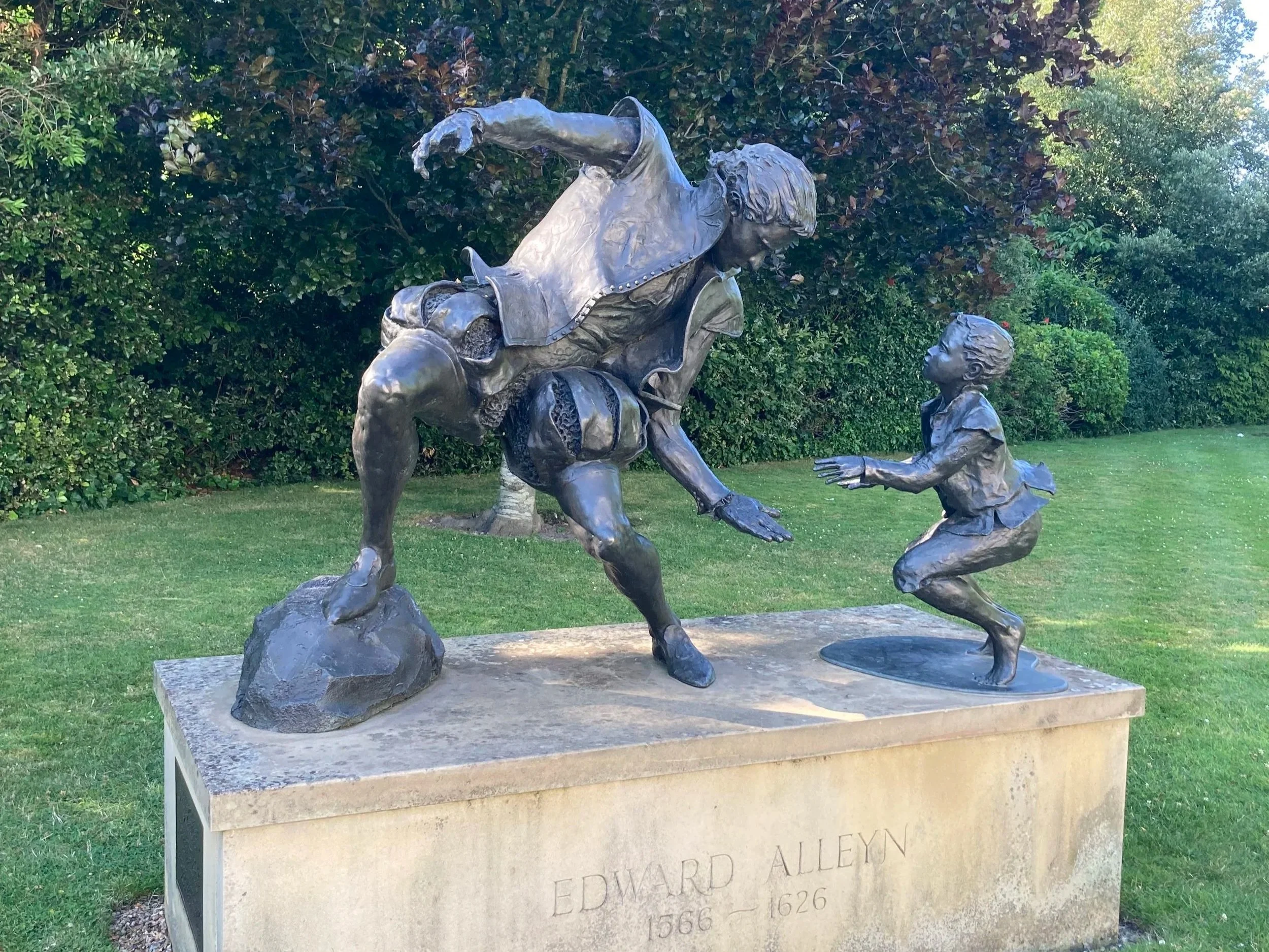 A bronze sculpture depicts a man with long hair, standing on the ground, and a young boy kneeling and reaching out to the man. The man appears to be volunteering or presenting something to the boy. The sculpture is set outdoors against a background of dense green foliage and trees, mounted on a rectangular stone plaque with inscriptions.