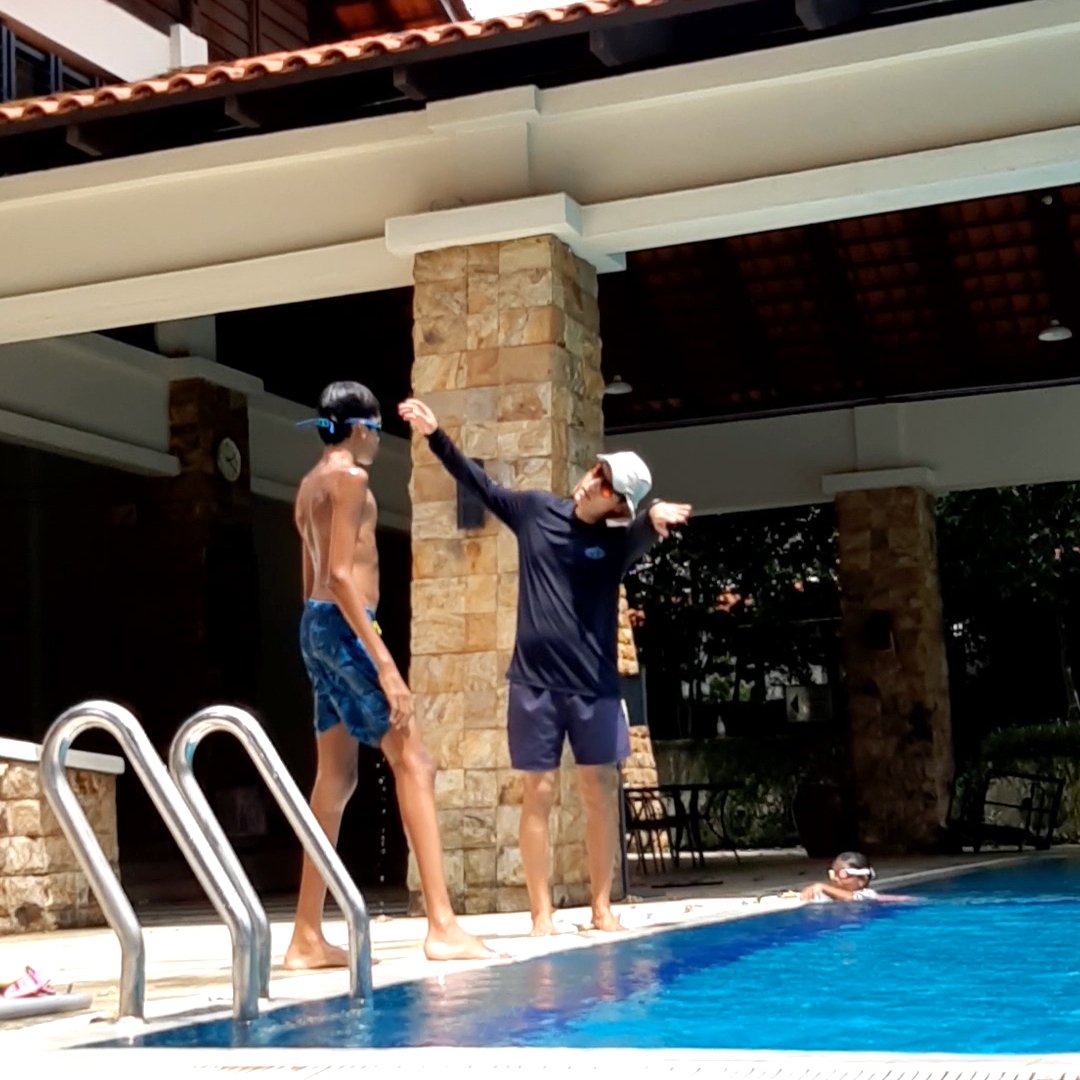 Two people standing at the edge of a swimming pool, one wearing goggles and swim shorts, the other in a hat and swimwear, gesturing with arms. Another person is visible swimming in the pool.