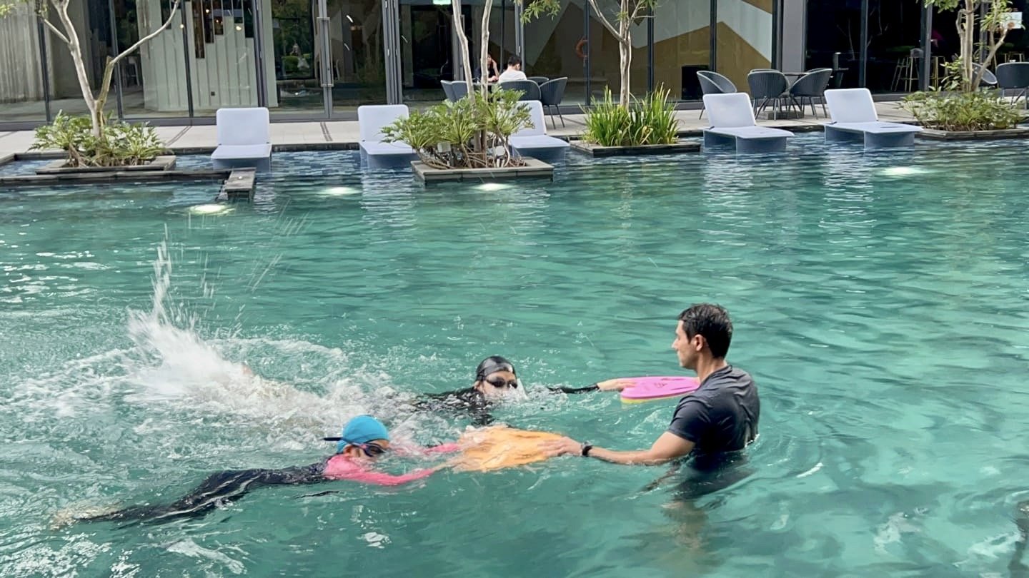 Children learning to swim in a pool with an instructor holding a swimming board, splashing water visible.