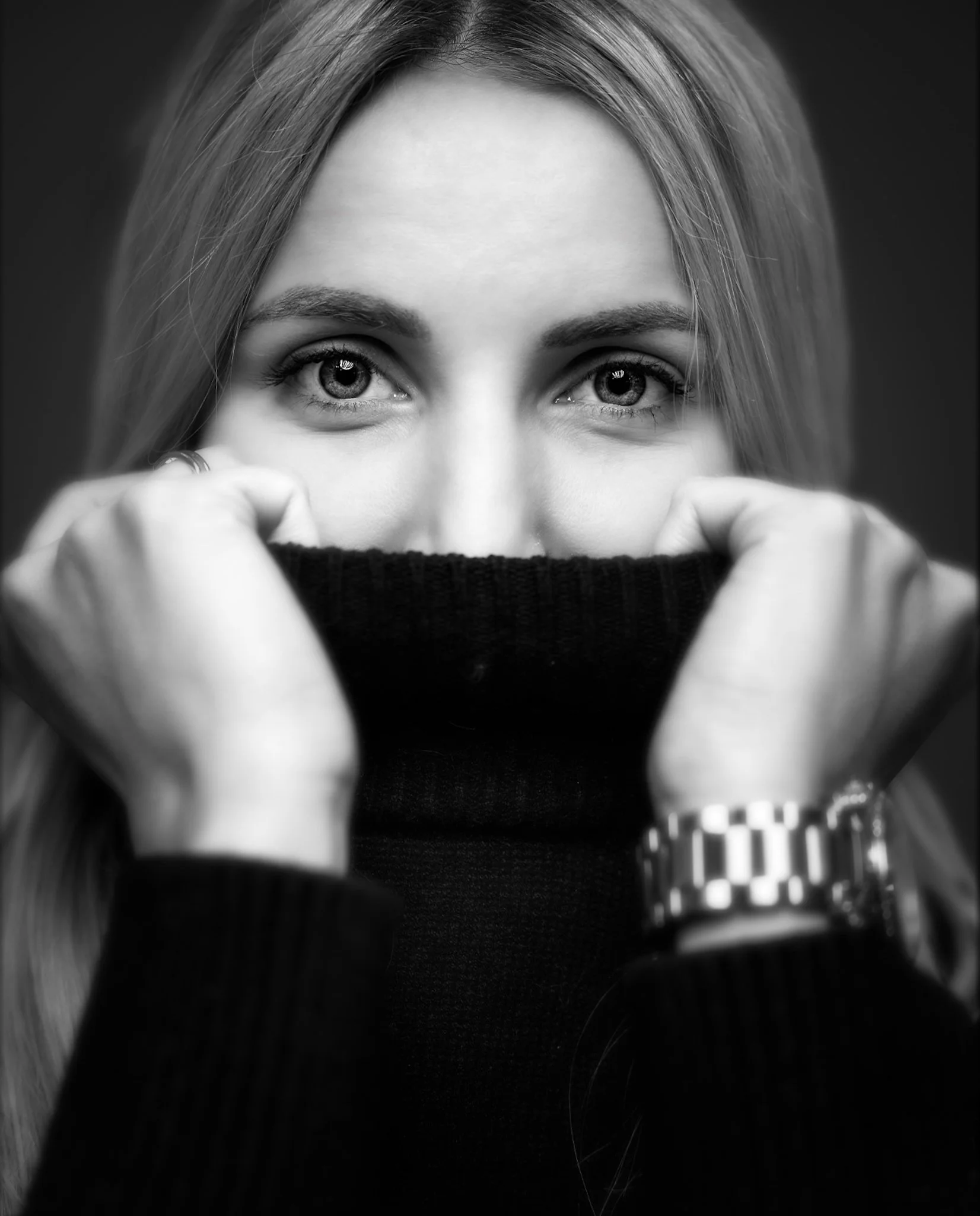 Schwarz-weiß-Fotografie einer jungen Frau, die ihr schwarzes Pullover bis zur Nase hochzieht, sodass nur ihre leuchtenden Augen sichtbar sind, während sie direkt in die Kamera blickt.