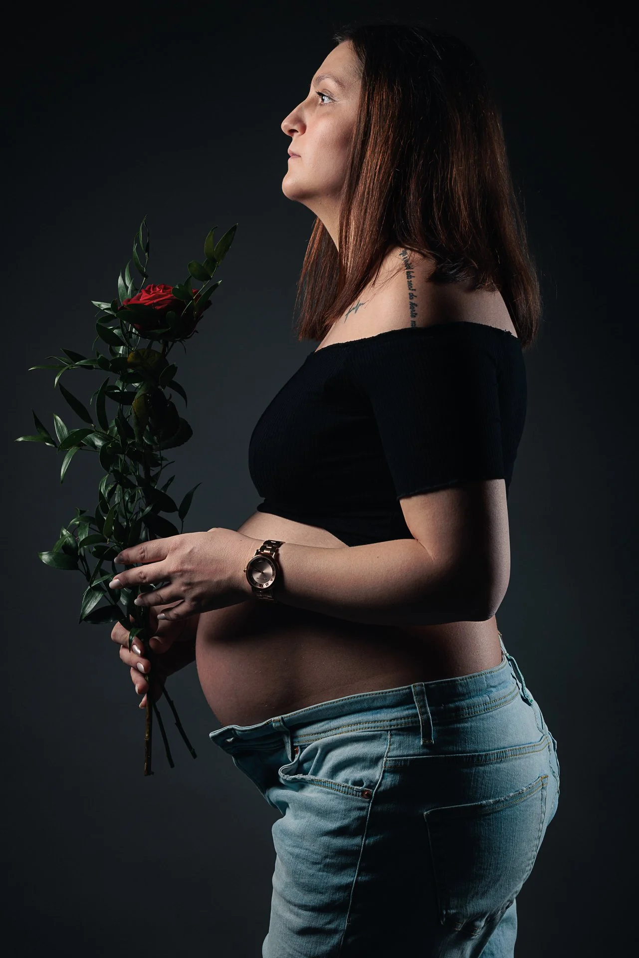Eine schwangere Frau mit braunen Haaren, die eine Blume in der Hand hält, trägt eine schwarze Oberteil und eine Uhr am Handgelenk, vor dunklem Hintergrund.