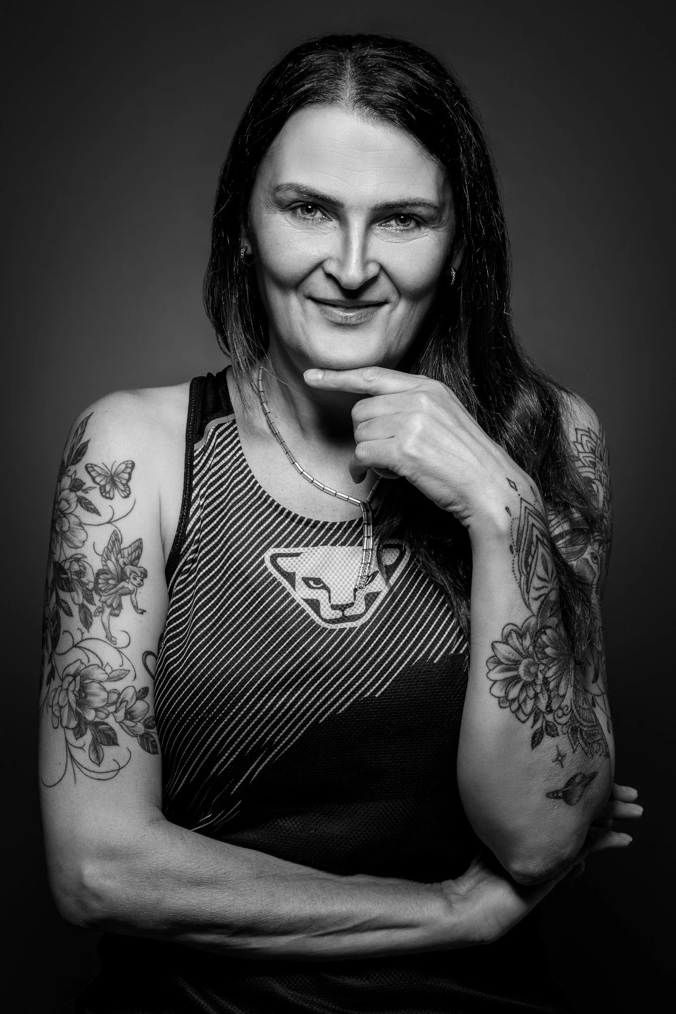 Schwarz-Weiß-Porträt einer lächelnden Frau mit Tattoos auf Armen und Schulter, trägt ein Sporttop mit Puma-Logo und ein Kettenhalsband.