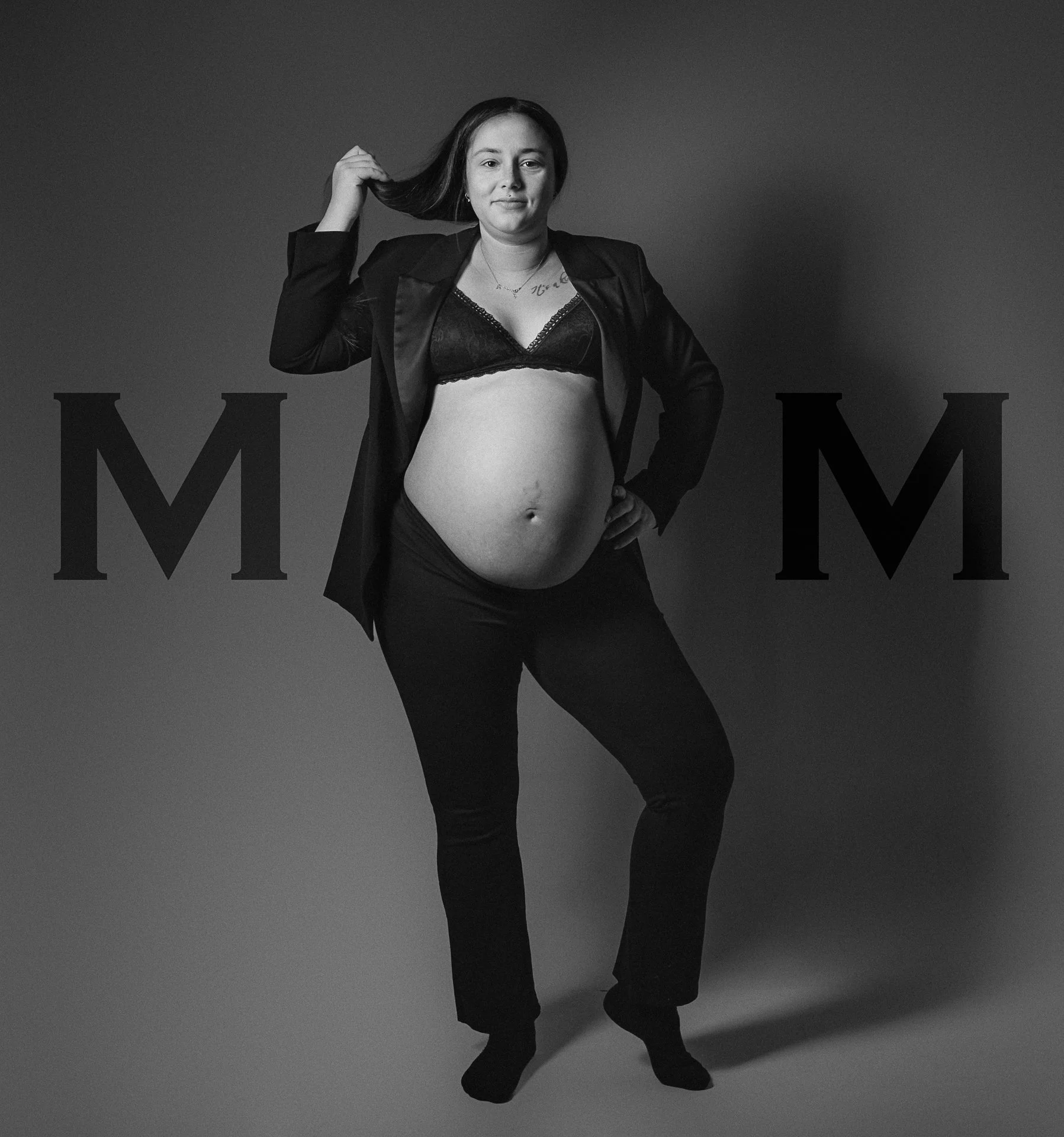 Schwangere Frau posiert für ein schwarz-weiß-Foto, trägt schwarze Kleidung und eine schwarze Jacke, steht vor einem grauen Hintergrund, mit großen Buchstaben M auf beiden Seiten.