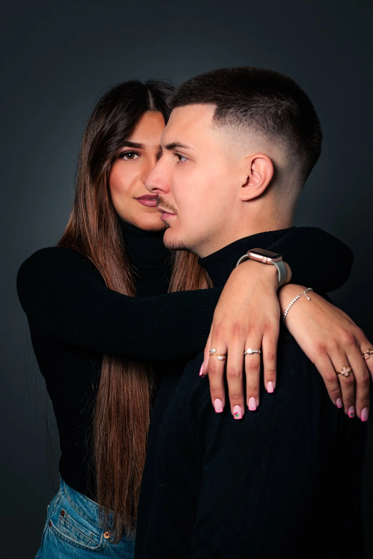 Ein Paar umarmt sich, die Frau schaut direkt in die Kamera, der Mann blickt seitlich, beide tragen schwarze Kleidung, die Frau hat lange braune Haare und trägt Schmuck, sie stehen vor dunkelgrünem Hintergrund. Fine-Art Fotografie mitten in den Alpen.