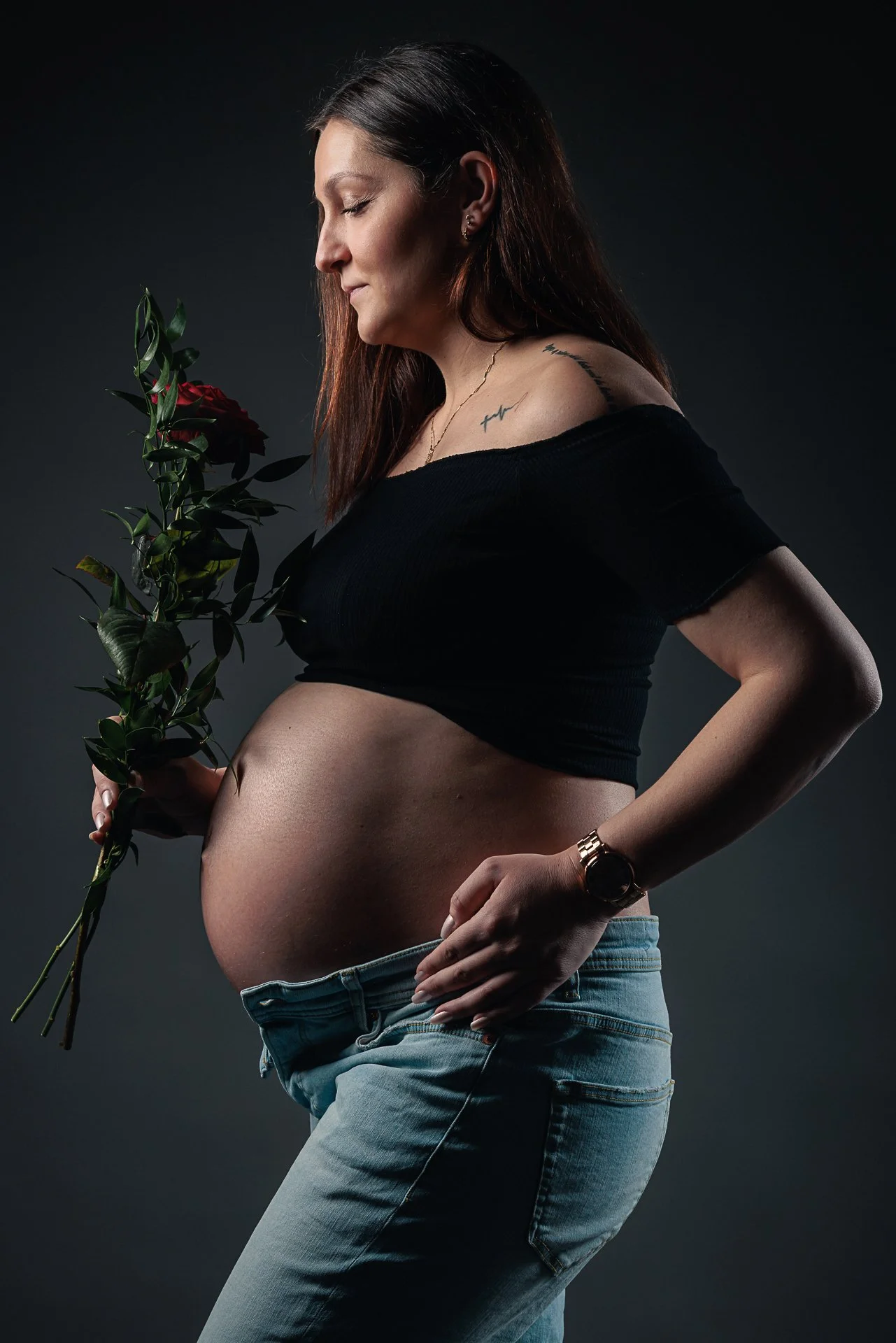 Eine schwangere Frau hält eine rote Rose vor sich und blickt liebevoll auf ihren Bauch. Sie trägt ein schwarzes off-the-shoulder Oberteil, Jeans und eine Armbanduhr.