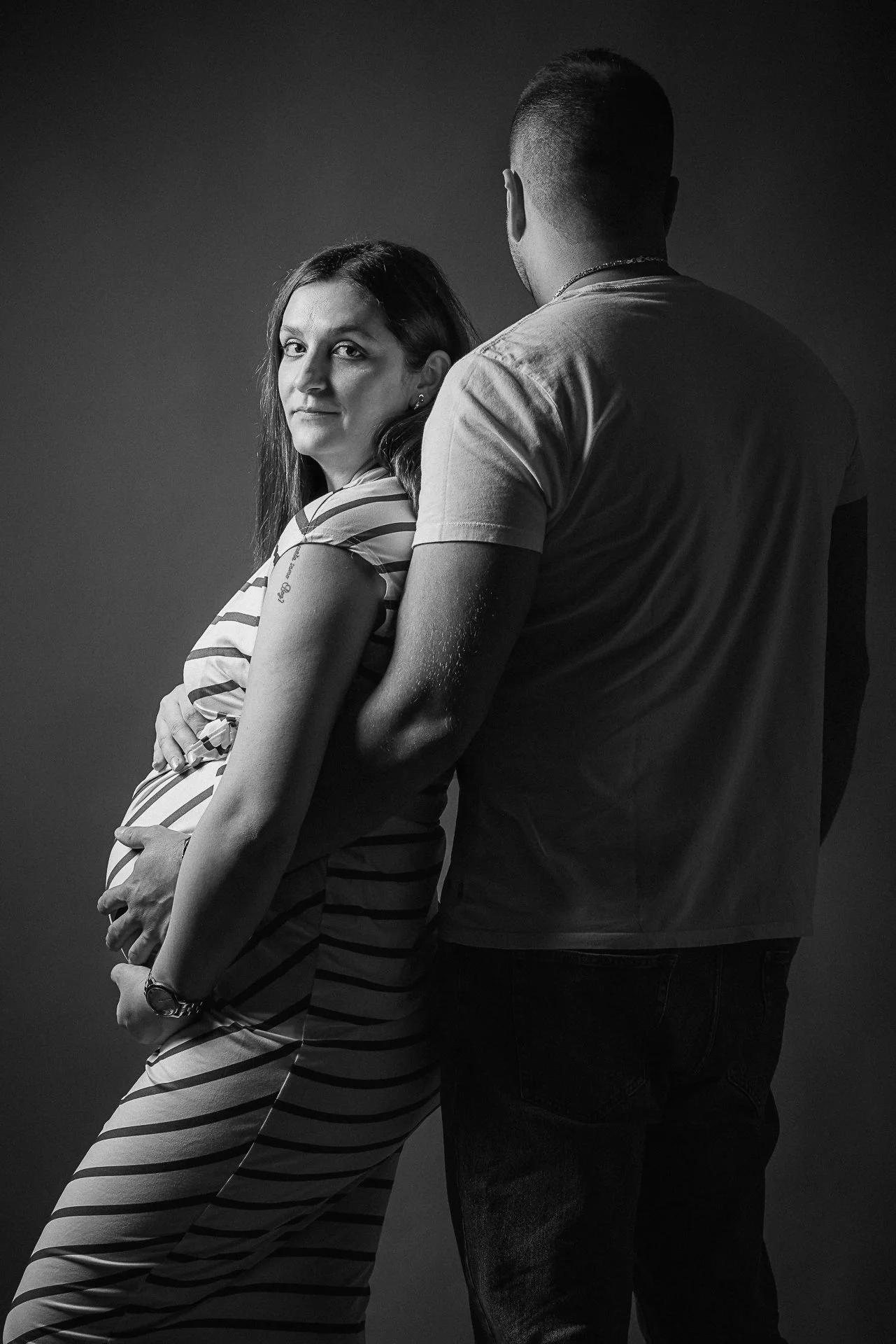 Schwangere Frau in gestreiftem Kleid wird von einem Mann gestützt, dunkler Hintergrund, Schwarz-Weiß-Fotografie.