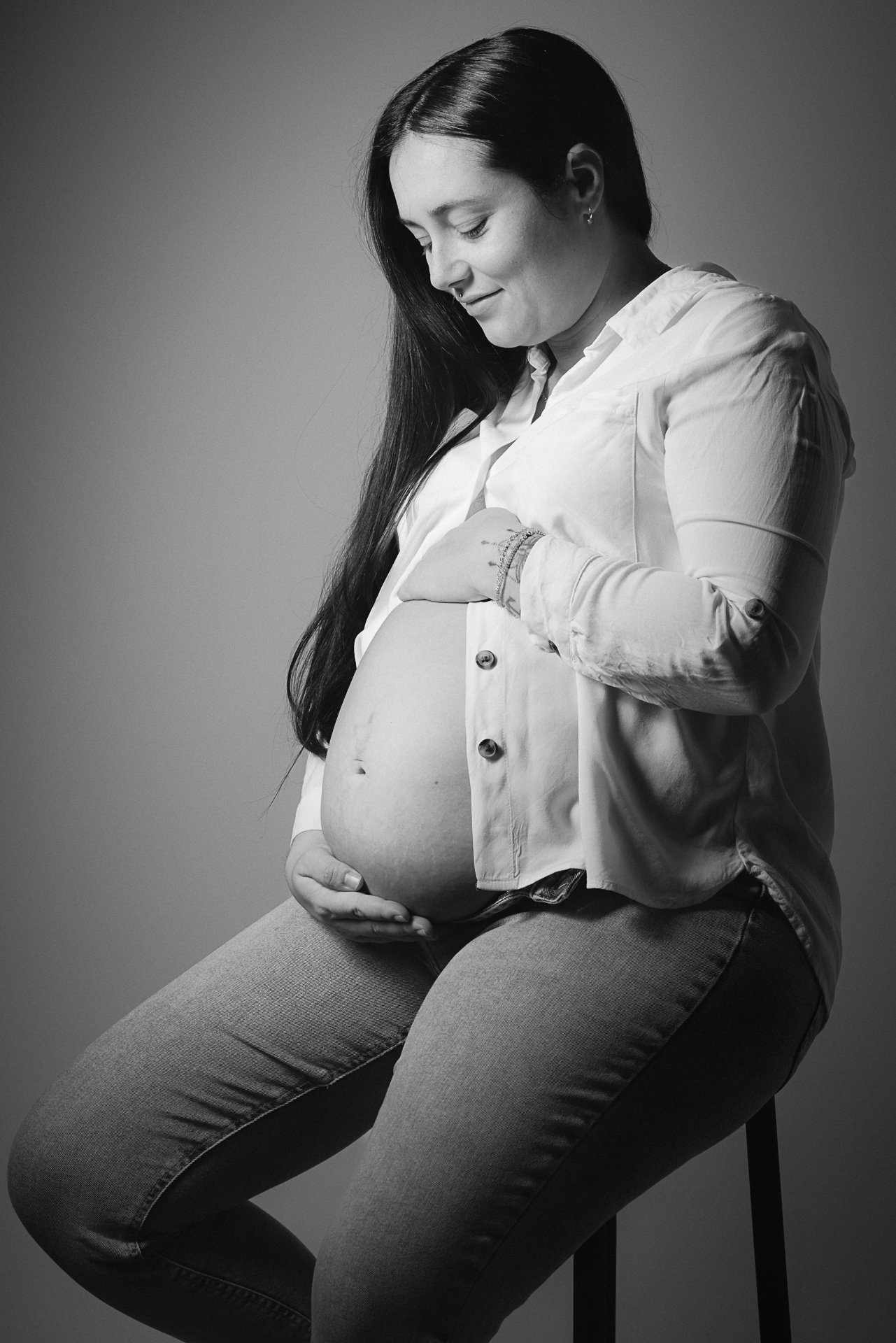 Schwangere Frau sitzt auf einem Stuhl, lächelt und schaut auf ihren Bauch, in Schwarz-Weiß-Fotografie.