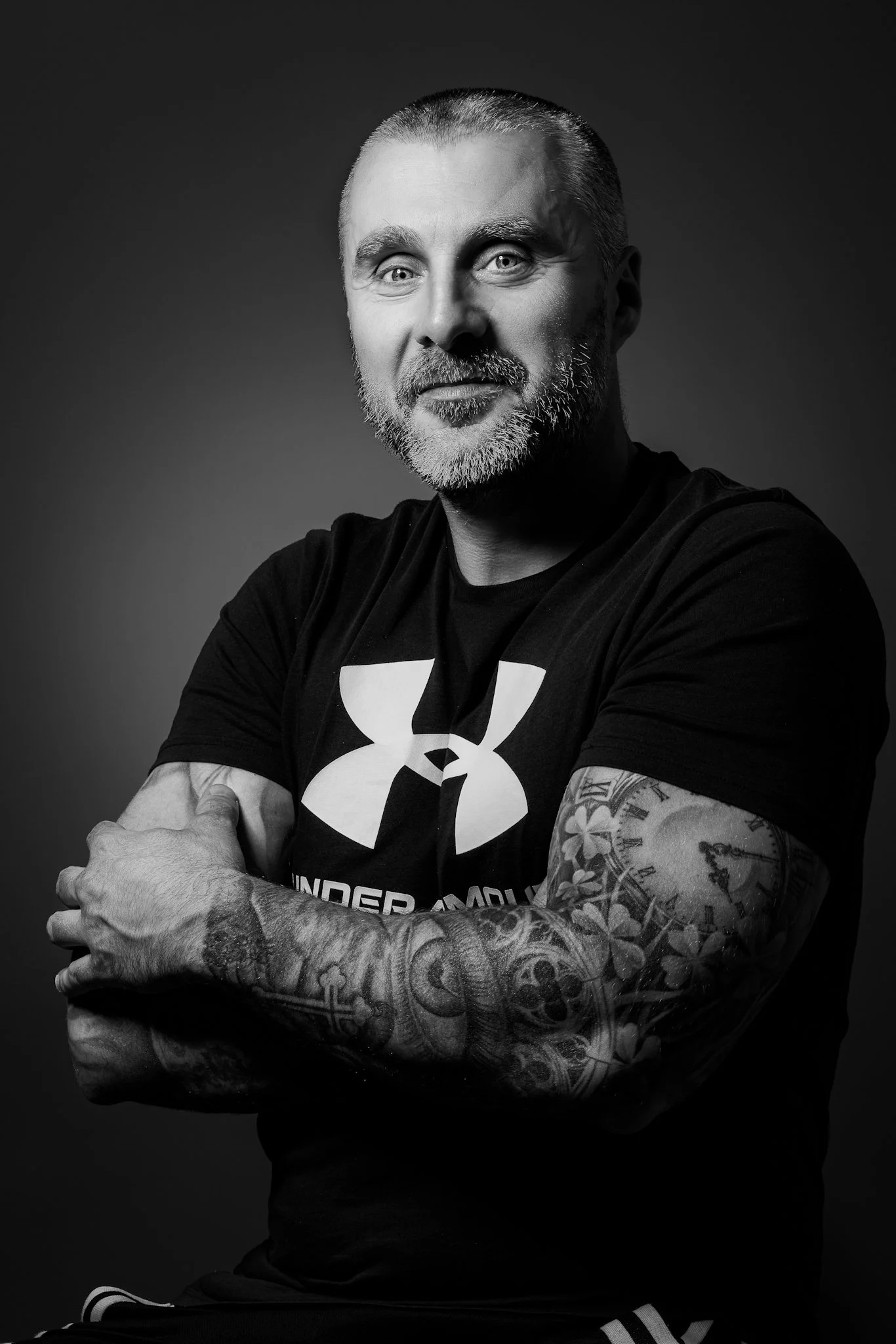 Schwarz-weiß Portrait eines Mannes mit tätowierten Armen, kurzen Haaren und einem Bart, trägt ein Under Armour T-Shirt, schaut in die Kamera, lächelt leicht und steht vor einem dunklen Hintergrund.