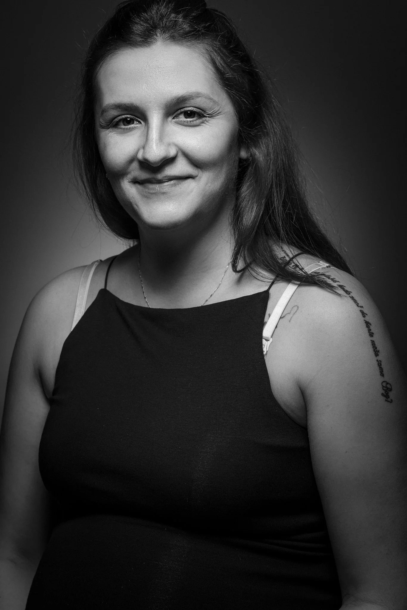Schwarz-weiß-Porträt einer lächelnden Frau mit dunklen Haaren, Tattoos am Schulterarm, trägt ein schwarzes Top mit dünnen Trägern, Hintergrund dunkel.