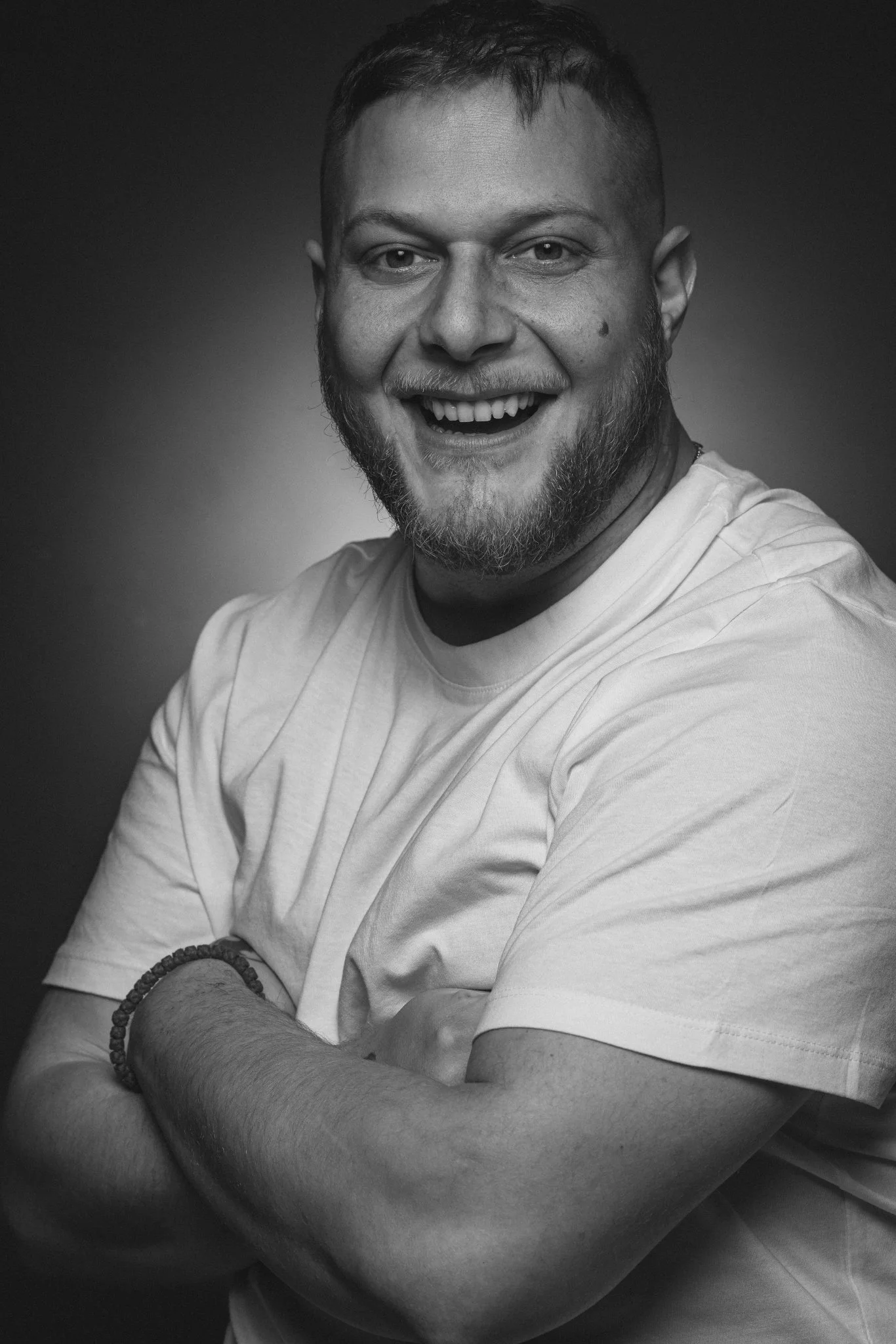 Schwarzweiß-Foto eines lachenden jungen Mannes mit Bart, der ein weißes T-Shirt trägt und die Arme verschränkt.