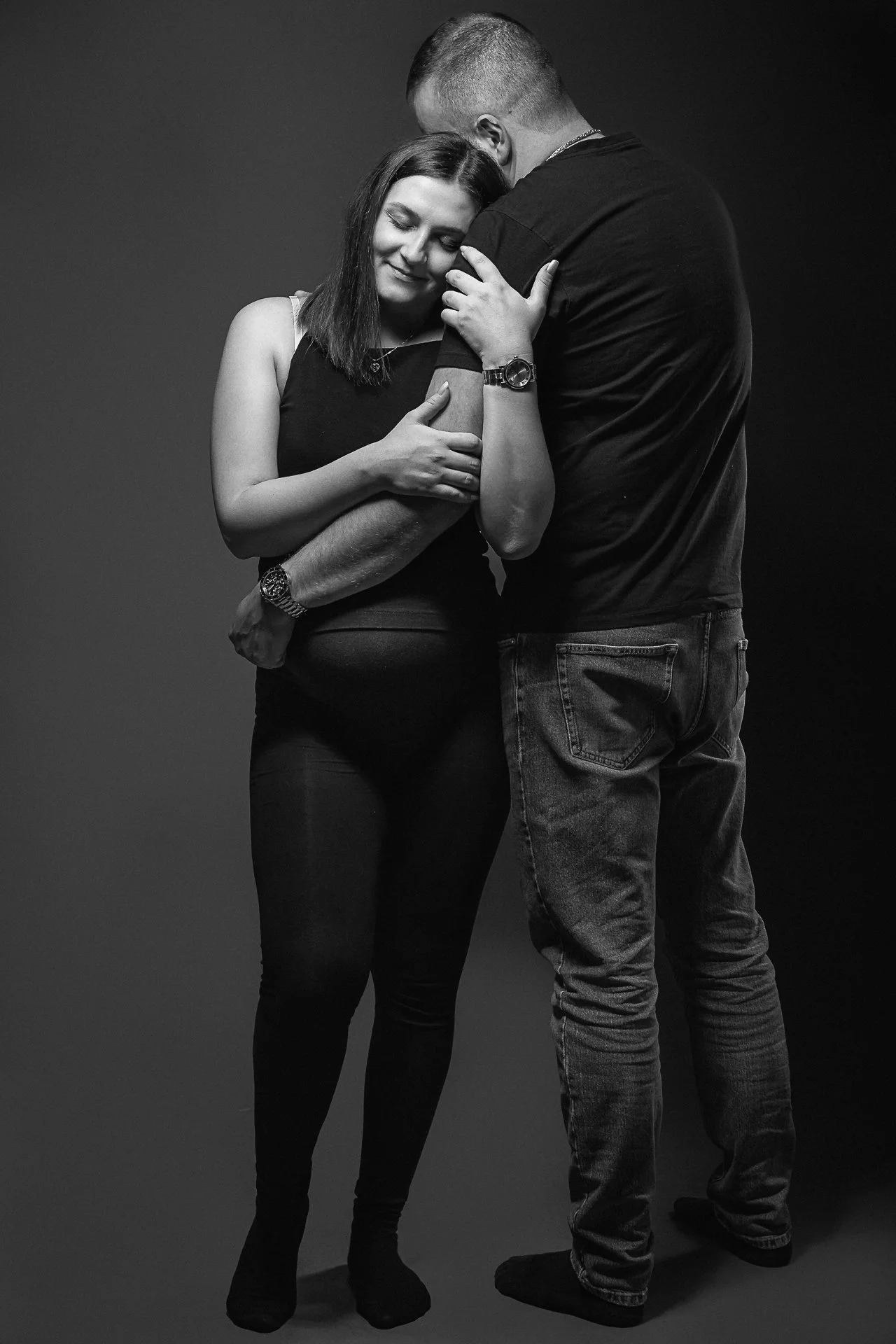 Ein Paar umarmt sich innig vor dunklem Hintergrund, die Frau trägt schwarze Kleidung, das Paar zeigt Zuneigung.