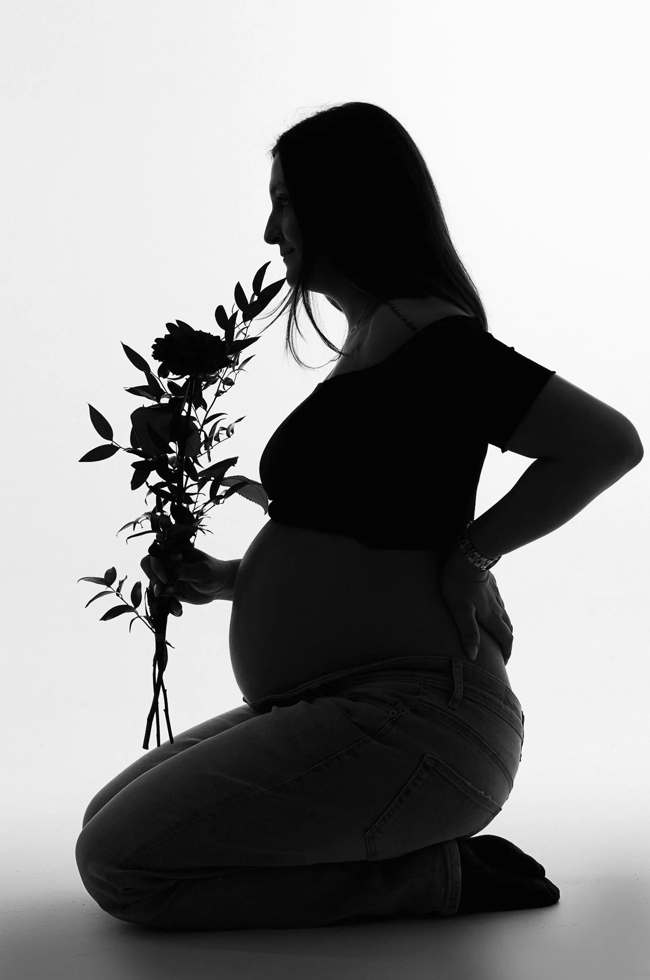 Silhouette einer schwangeren Frau, die auf den Knien sitzt, eine Blume hält und an einem weißen Hintergrund steht.