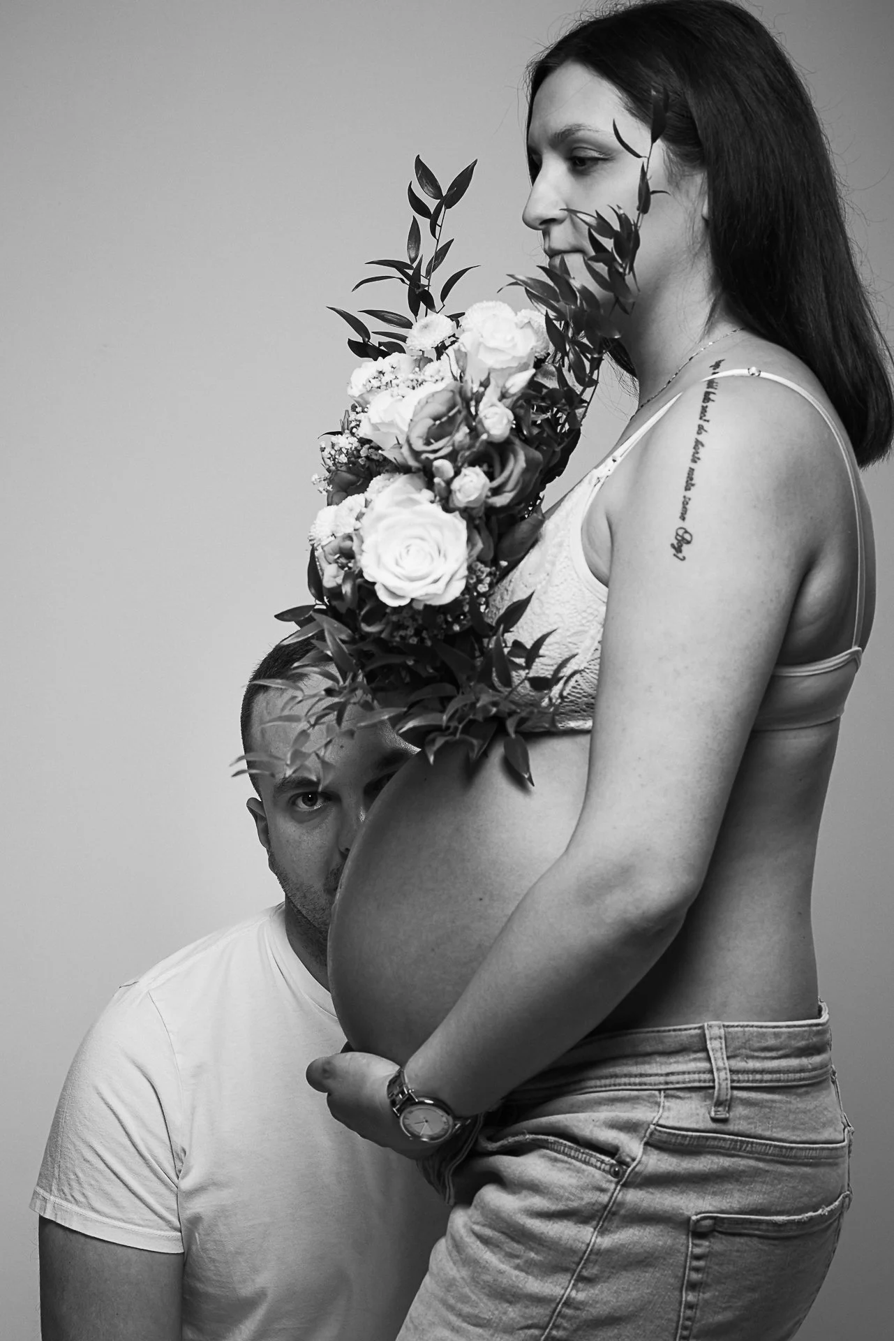 Schwangere Frau mit dunklen Haaren hält einen Blumenstrauß, während ein Mann hinter ihr halb sichtbar auf sie schaut.